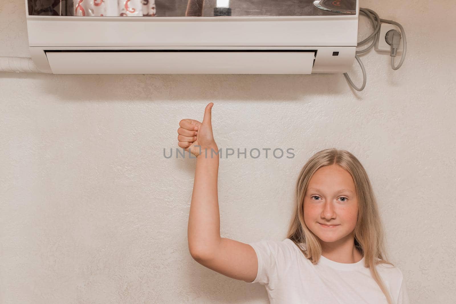 Young cute girl teenager blonde European appearance shows a cool thumb, looks into the camera and smiles next to the air conditioner on the wall in the room by AYDO8