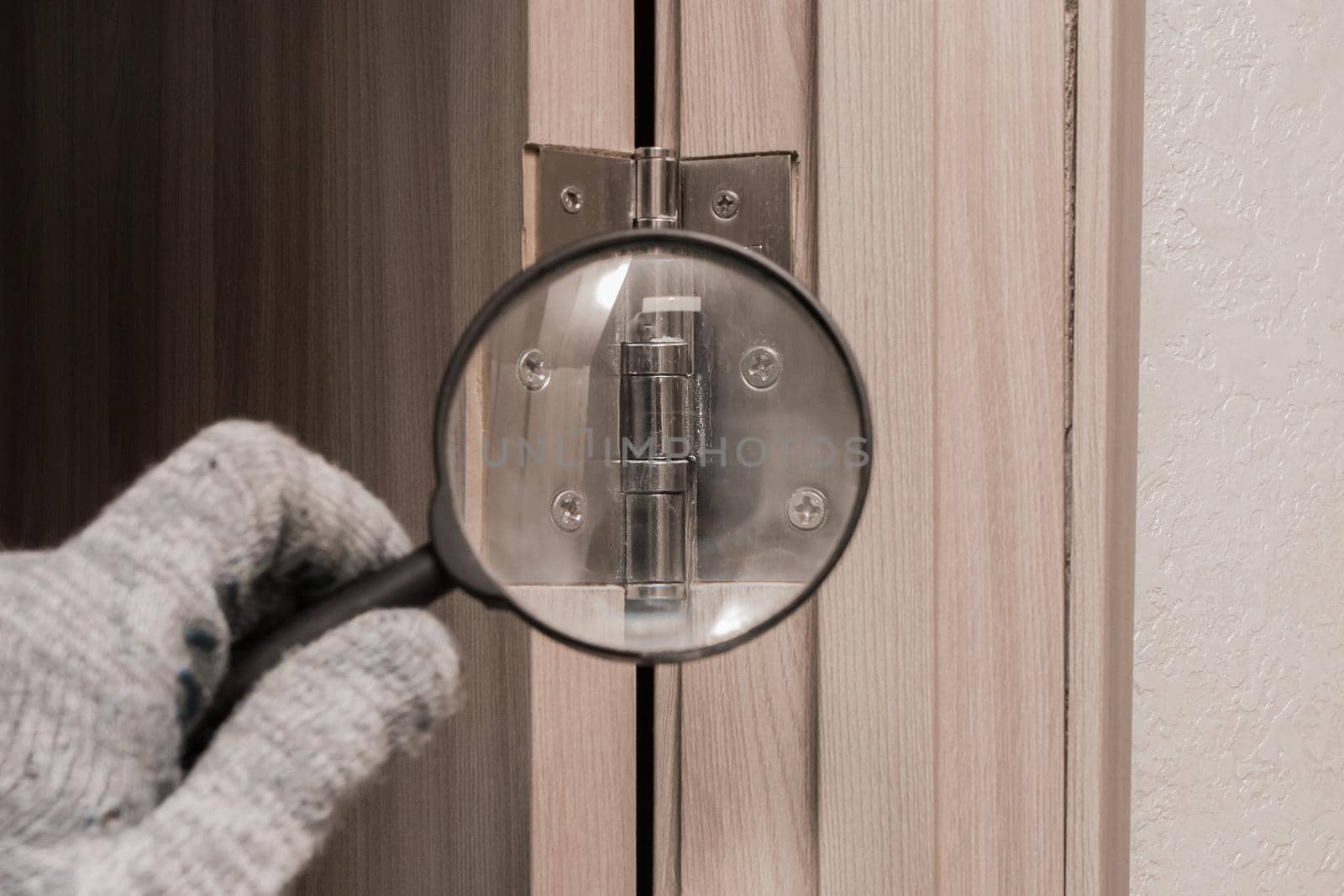 The hand of a male worker in a construction glove examines through a magnifying glass the door hinge of a wooden door. The concept of installation and fixation of metal parts.