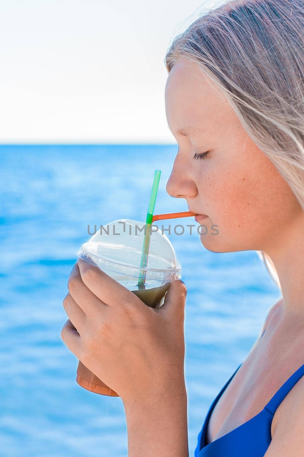 A young girl with blonde hair of European appearance, a teenager holds and drink a colored cold non-alcoholic cocktail in her hand against the background of the sea beach.