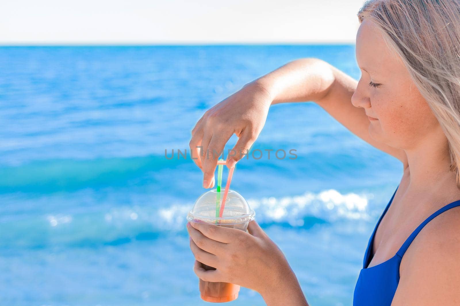 The hand of a young girl in a blue swimsuit holds a cooling, colored non-alcoholic cocktail against the background of the water of the sea beach and the shore by AYDO8