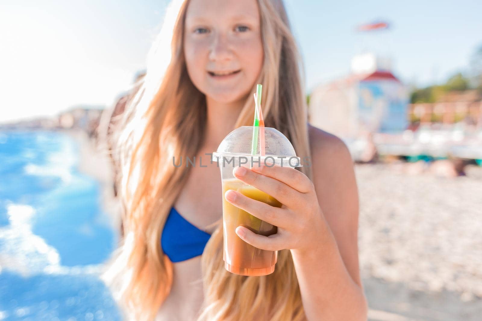 A young girl with blonde hair of European appearance, a teenager holds and drink a colored cold non-alcoholic cocktail in her hand against the background of the sea beach by AYDO8