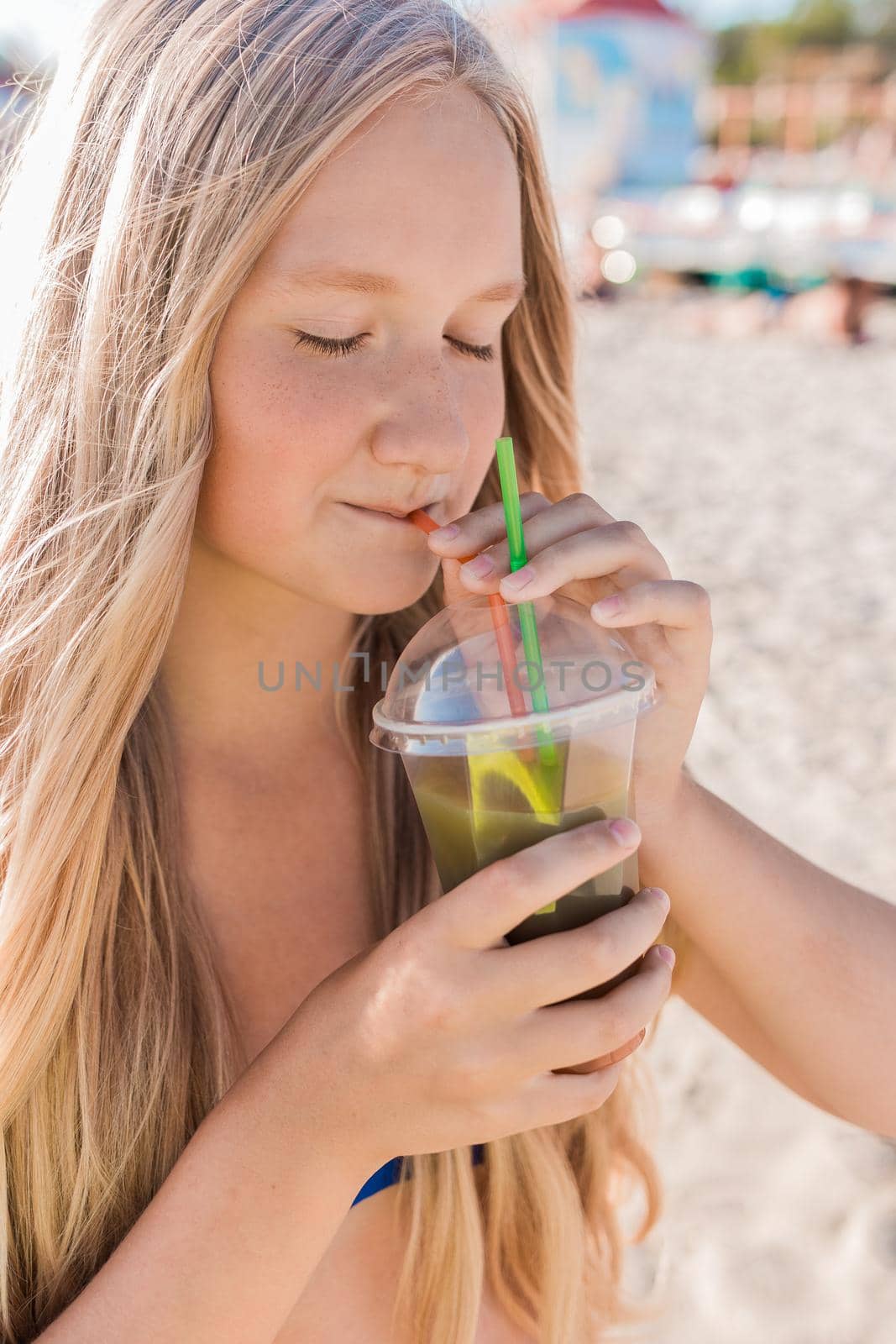 A young girl with blonde hair of European appearance, a teenager holds and drink a colored cold non-alcoholic cocktail in her hand against the background of the sea beach by AYDO8