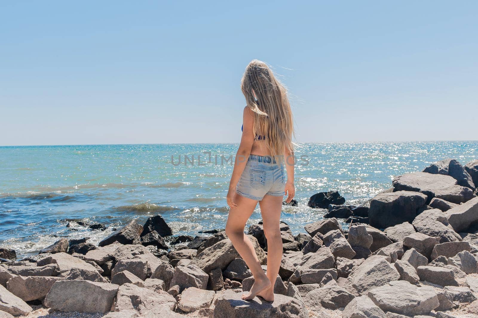 A young slender teenage girl with long blonde hair, in short denim shorts standing on the rocks of the breakwater on the beach near the seashore by AYDO8