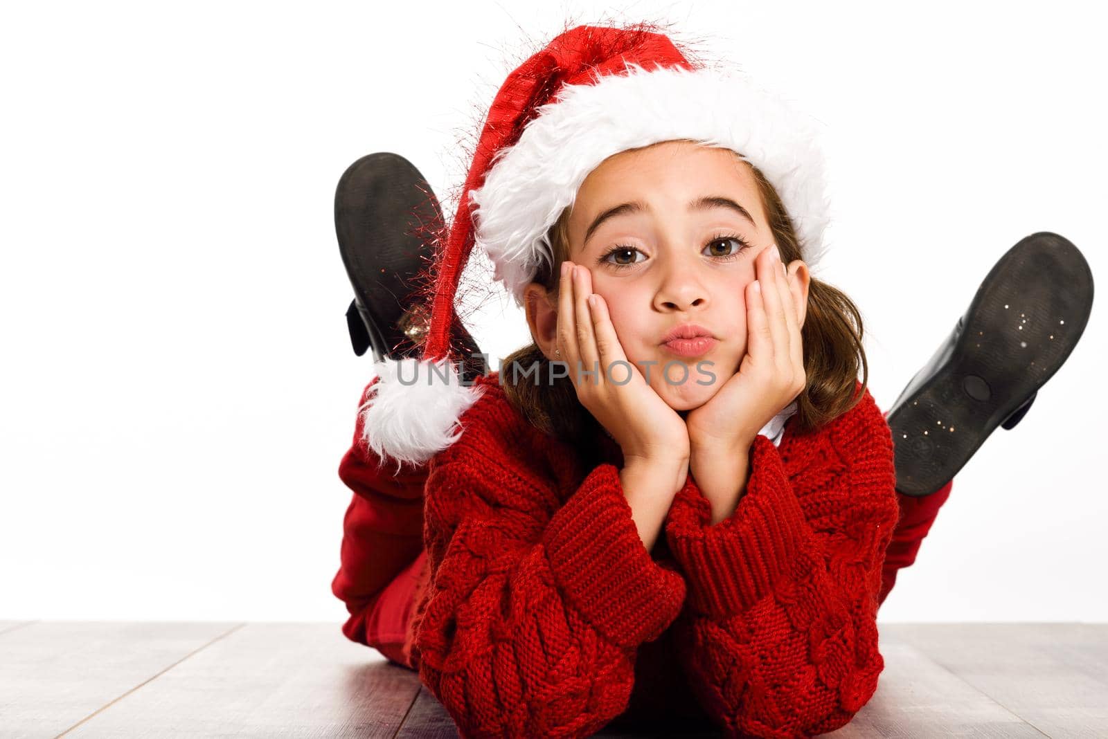 Adorable little girl wearing Santa hat laying on wooden floor on white background. Winter clothes