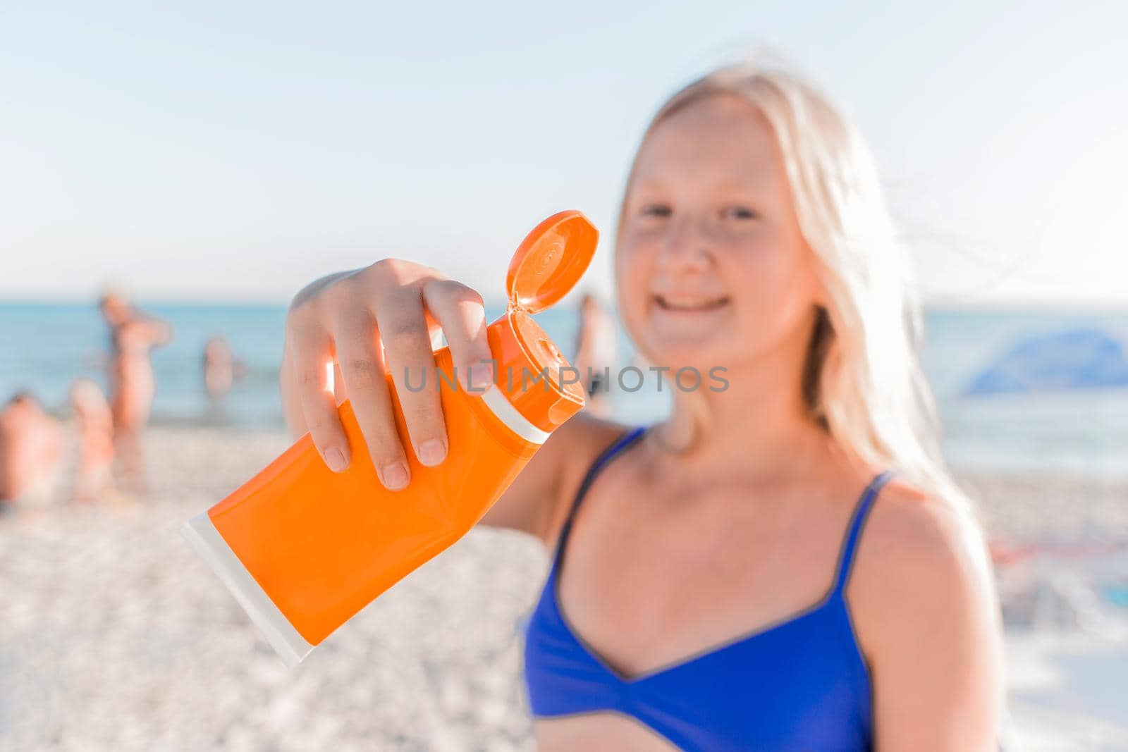 Young cute girl blonde teenager holding sunscreen in her hand against the background of the sea beach.