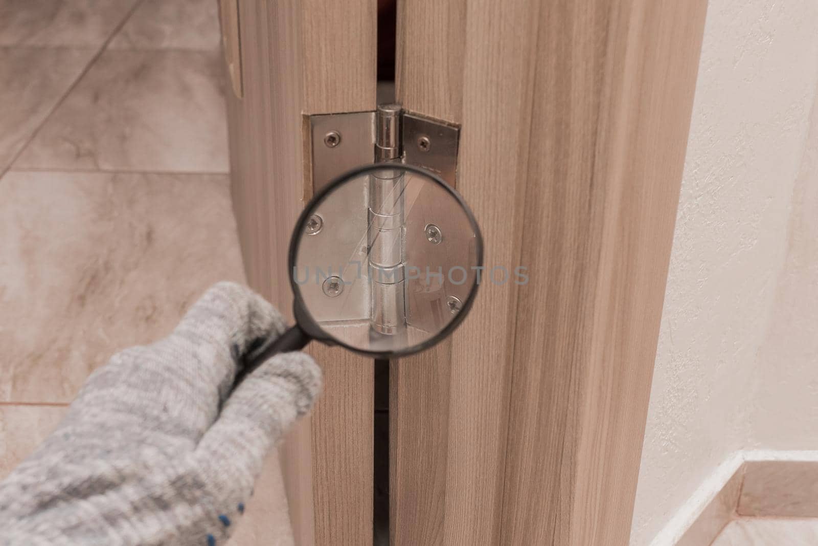 The hand of a male worker in a construction glove examines through a magnifying glass the door hinge of a wooden door. The concept of installation and fixation of metal parts by AYDO8