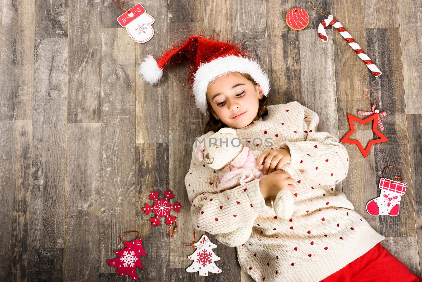 Adorable little girl wearing santa hat smiling on wooden floor with Christmas ornaments. Winter clothes.