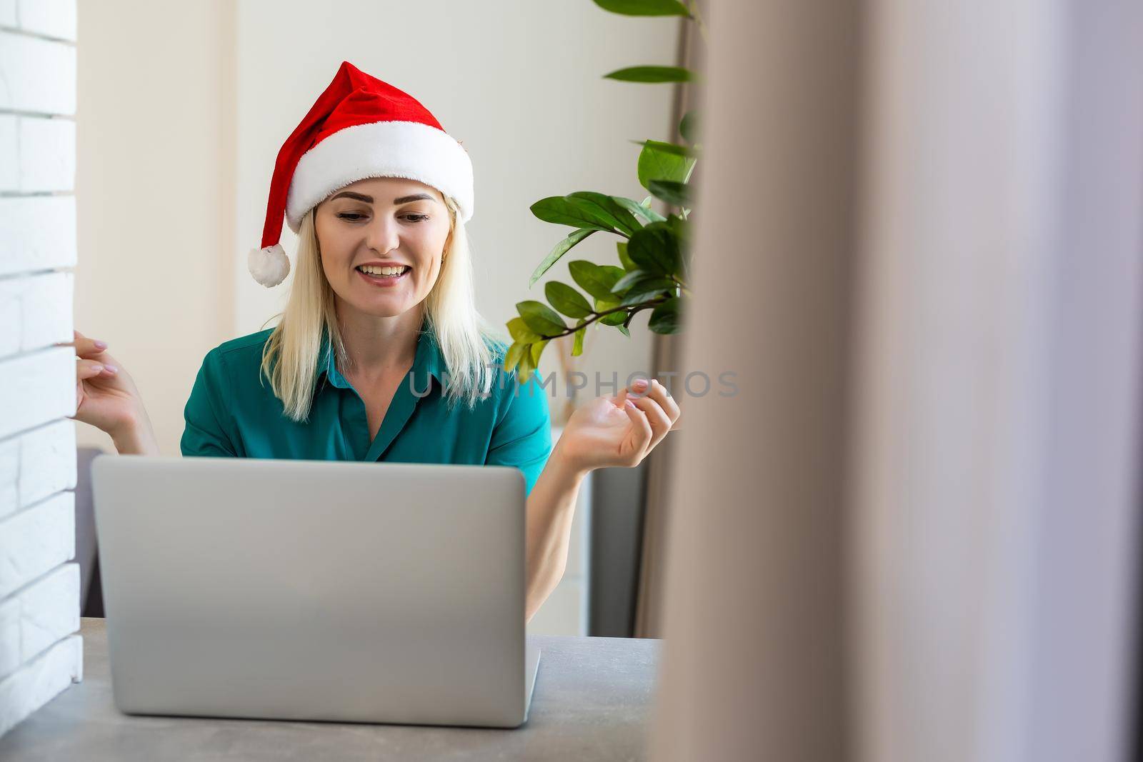 Woman in a red Santa hat communicates in a Christmas video conference. Girl celebrates Christmas at home during the pandemic. Communication with loved ones via the Internet. Online new year greetings