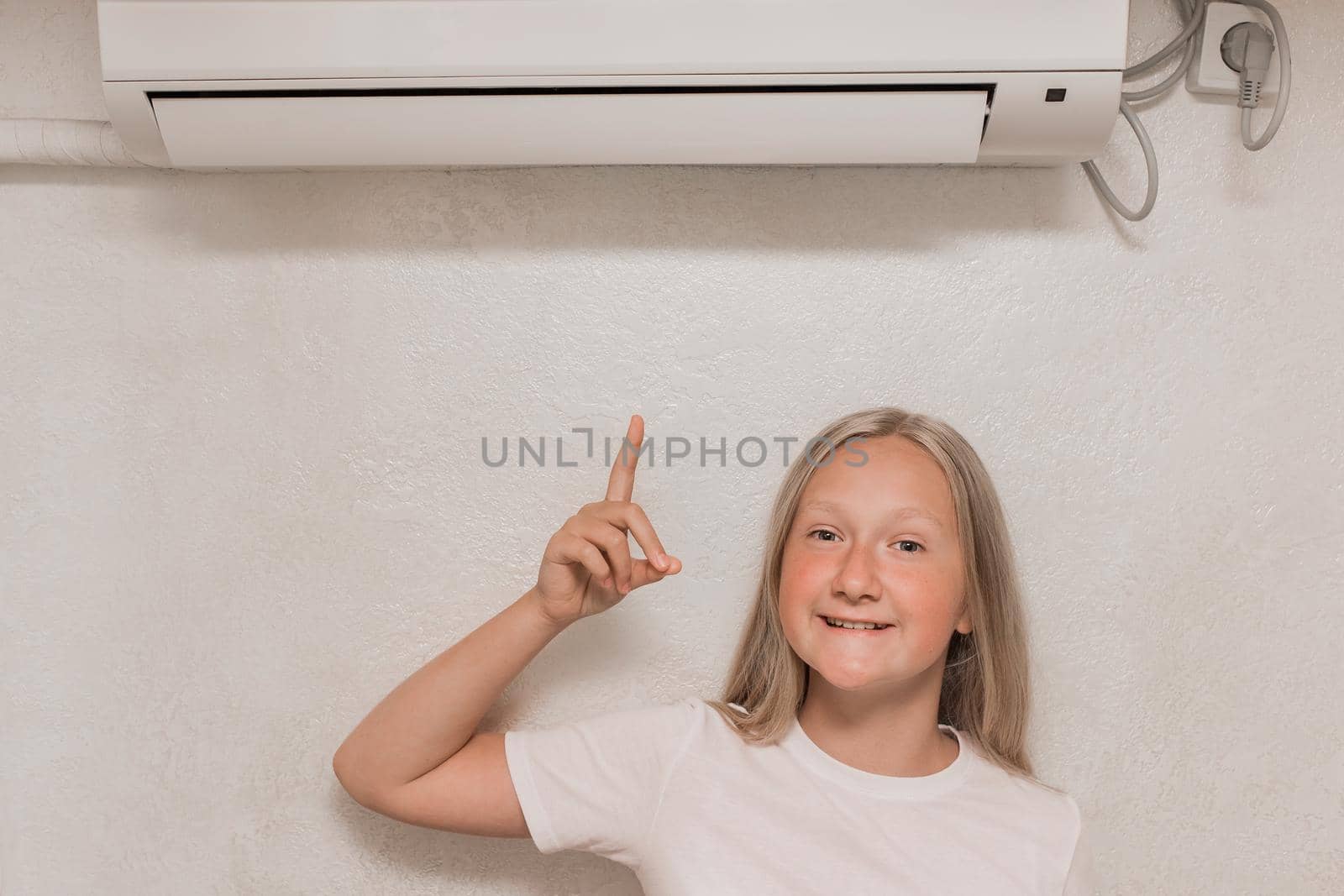Cute teenage girl blonde European appearance points a hand finger at the air conditioner on the wall in the room.
