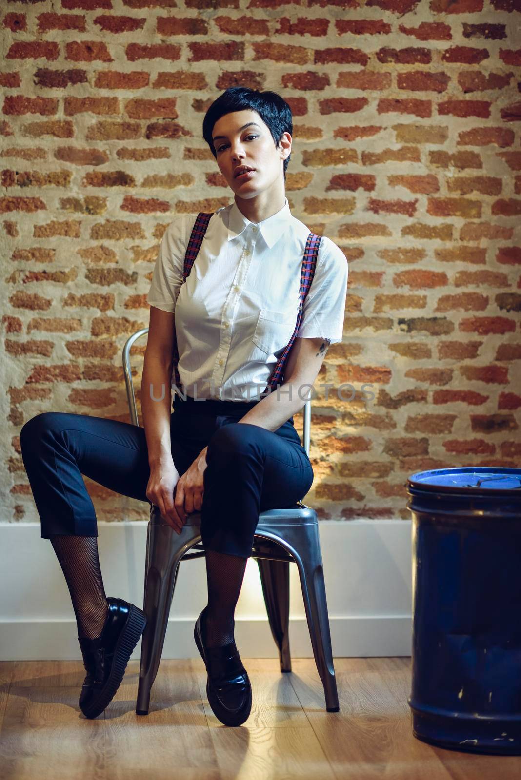 Beautiful young woman, model of fashion, with very short haircut, looking at camera sitting on chair, with a brick wall in the background. Beauty and hairstyles concept.