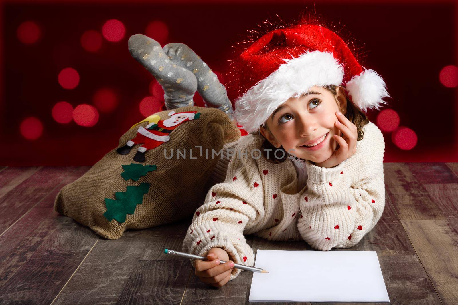 Adorable little girl wearing santa hat writing Santa letter on wooden floor. Winter clothes for Christmas. Red bokeh at the background