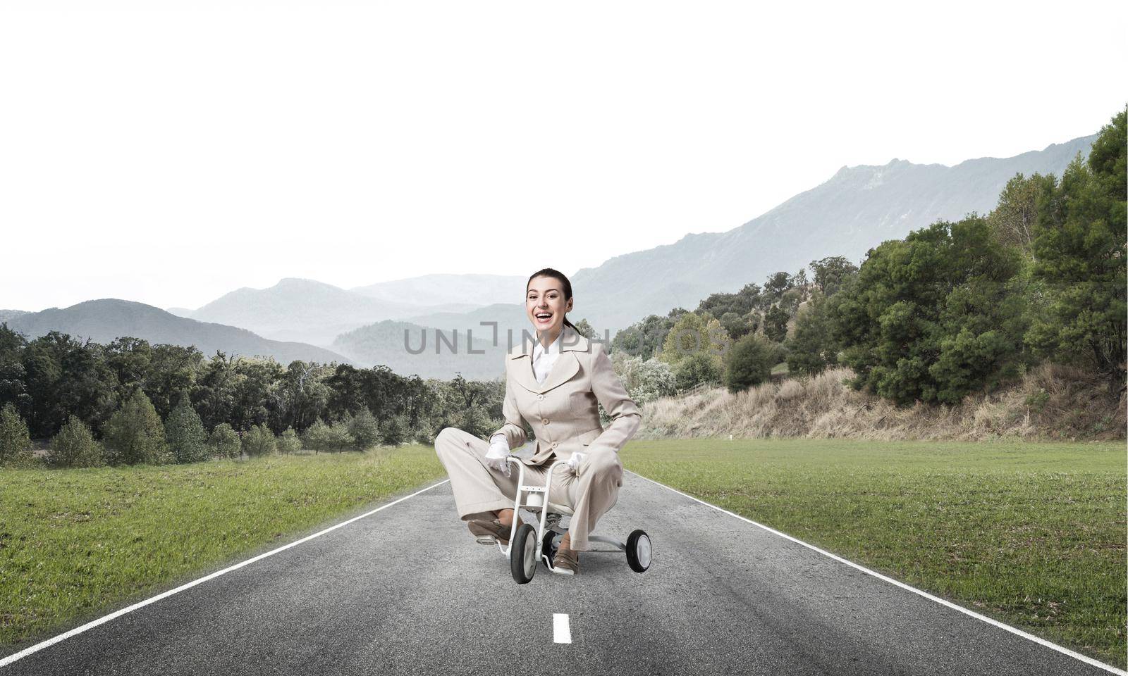 Beautiful happy woman riding children's bicycle on asphalt road. Young employee in white business suit cycling small bike outdoor. Nature landscape with green grass. Professional career start concept