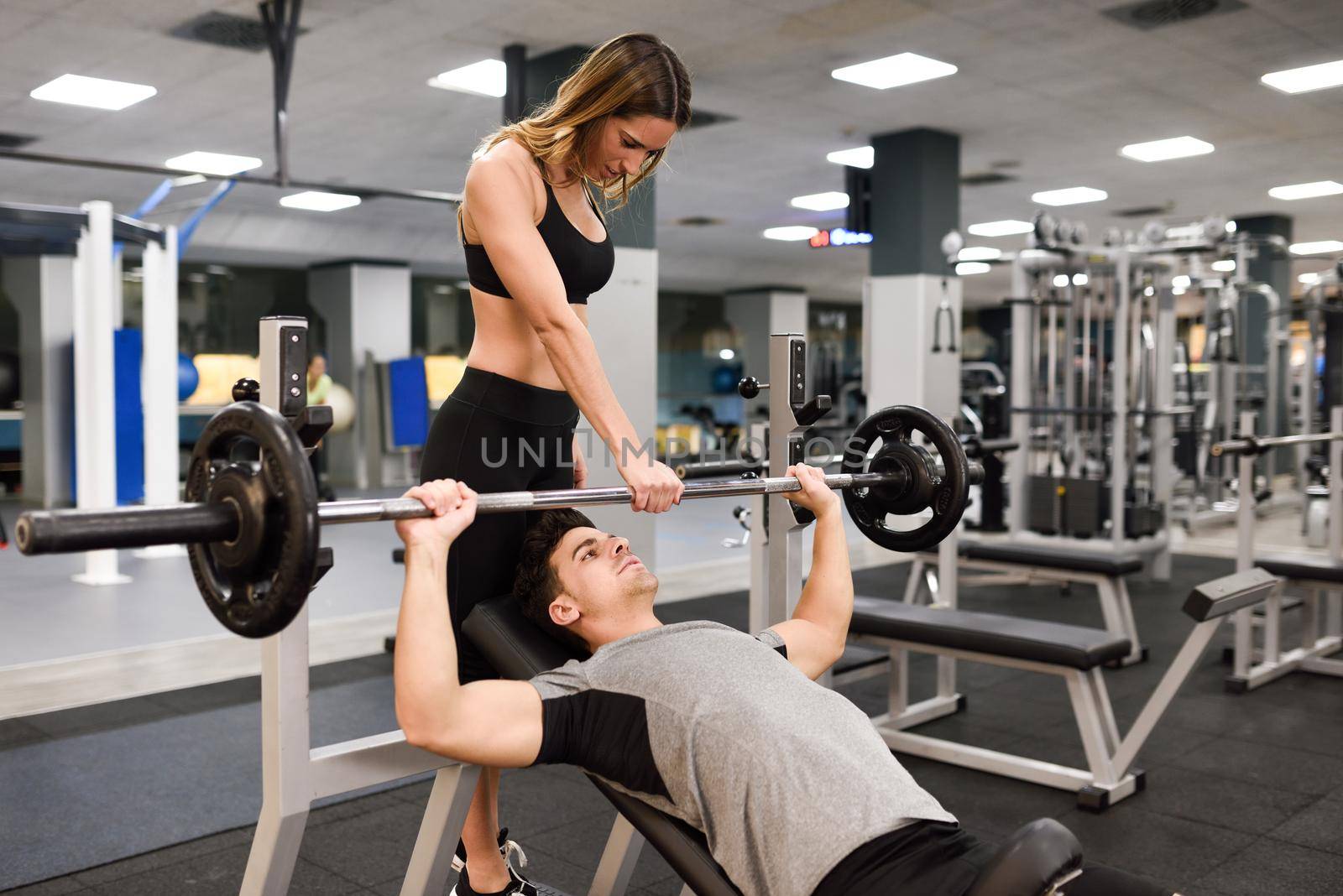 Female personal trainer helping a young man lift weights while working out in a gym