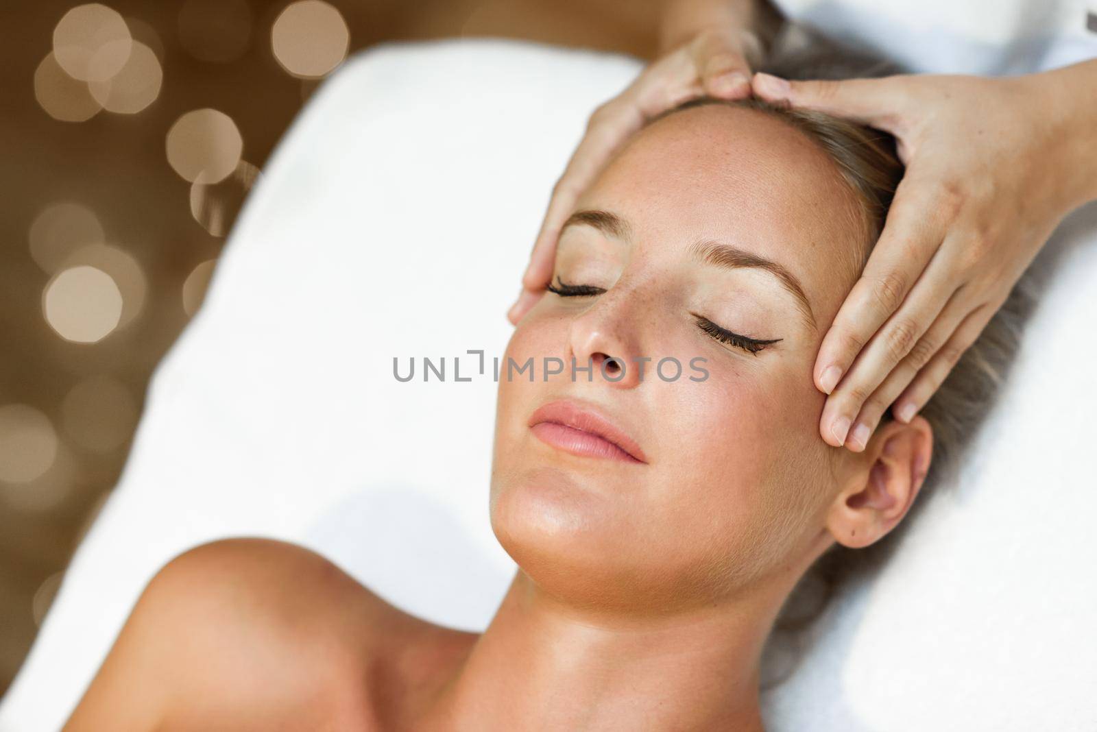 Young blond woman receiving a head massage in a spa center with eyes closed. Female patient is receiving treatment by professional therapist.