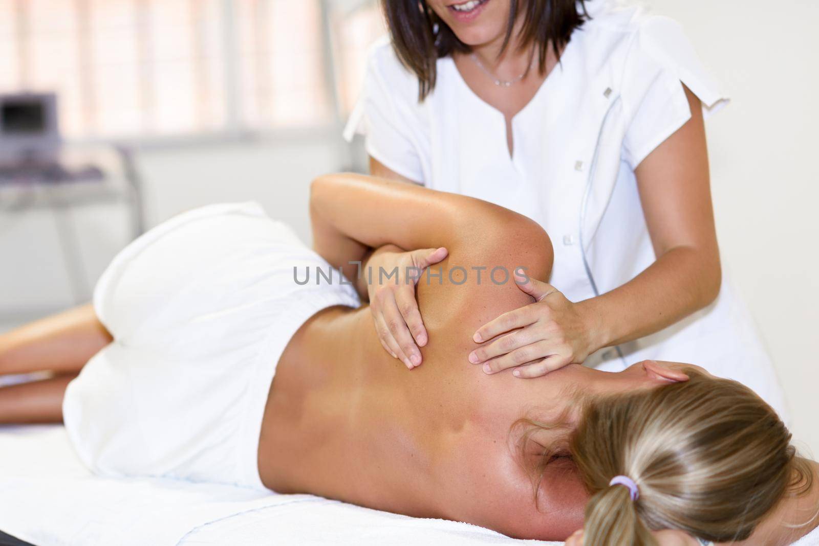 Professional female physiotherapist giving shoulder massage to blonde woman in hospital. Medical check at the shoulder in a physiotherapy center.