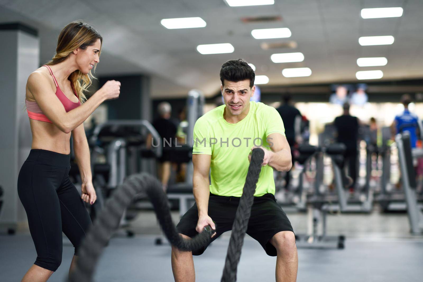 Female personal trainer motivating young man with battle ropes exercise in the fitness gym.