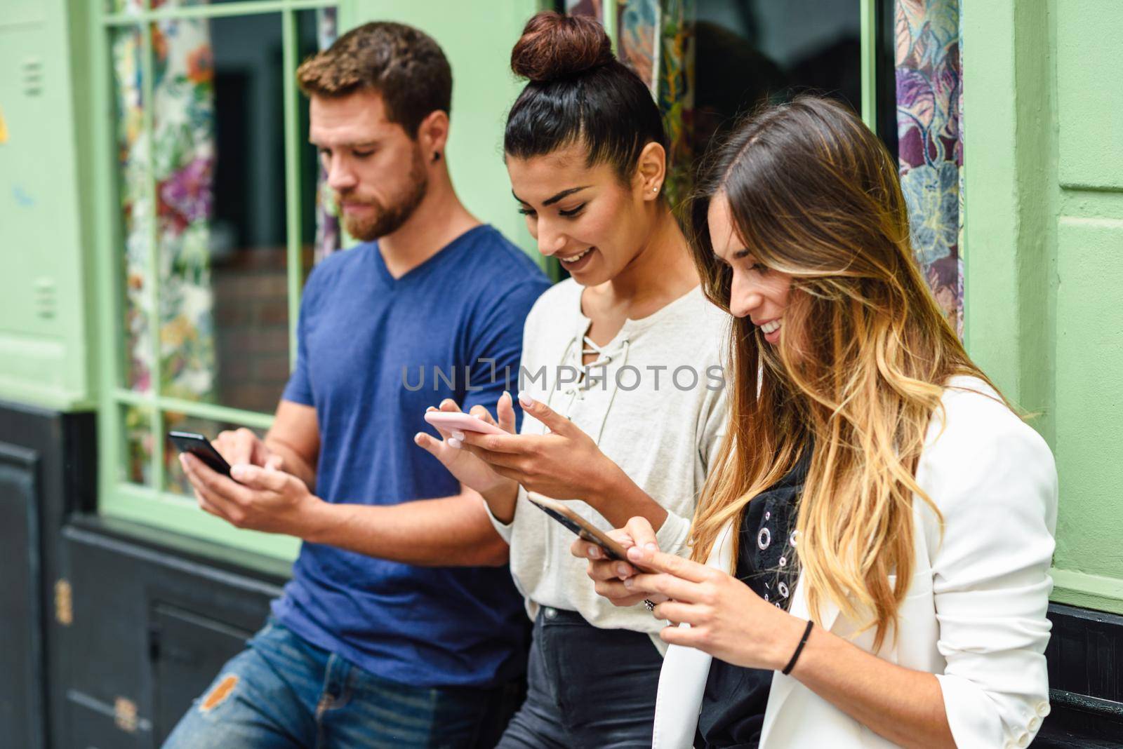 Multiethnic group of three people looking down at smart phone smiling, concepts about technology addiction and youth