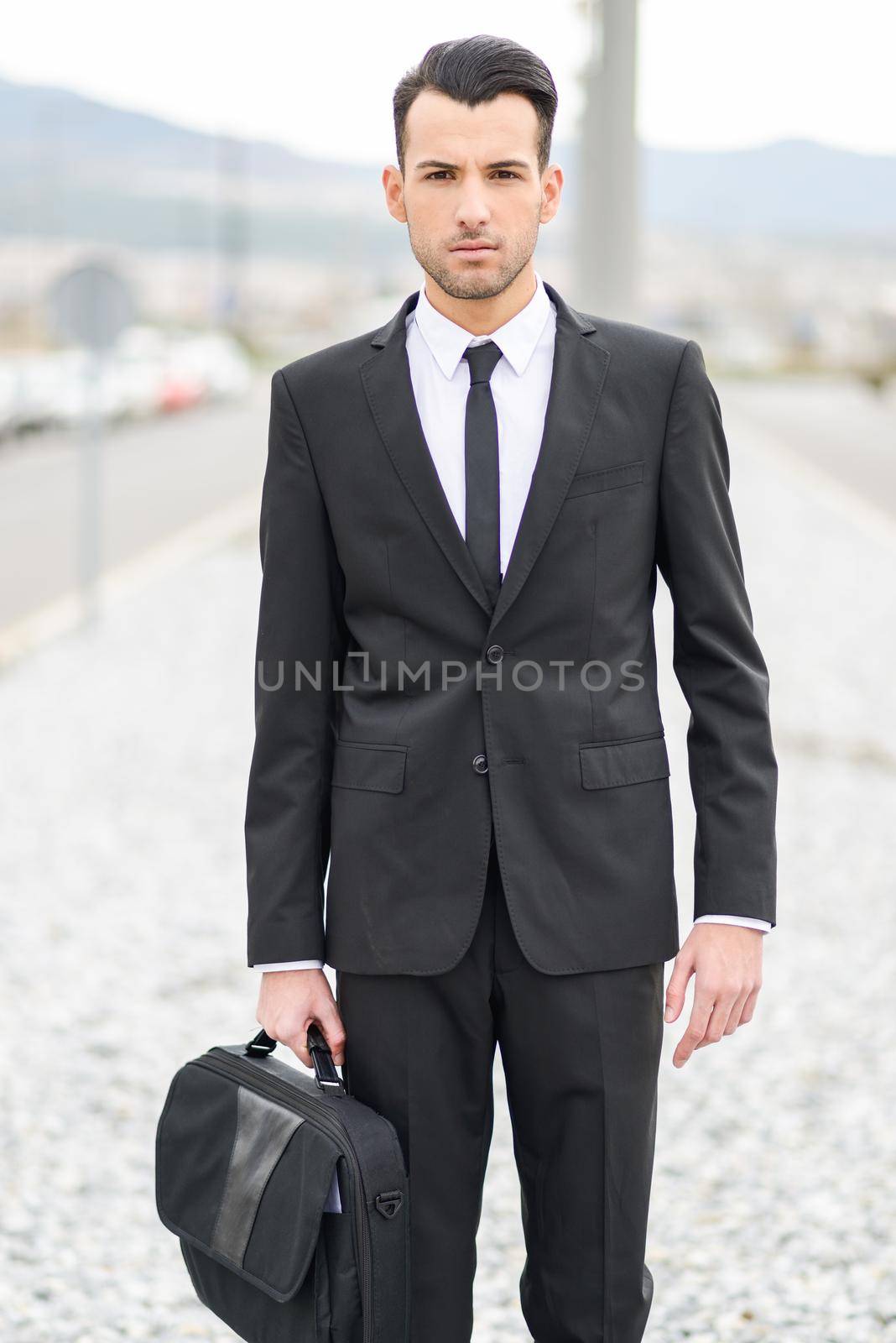 Portrait of a young businessman near a office building wearing black suit and tie with modern haircut