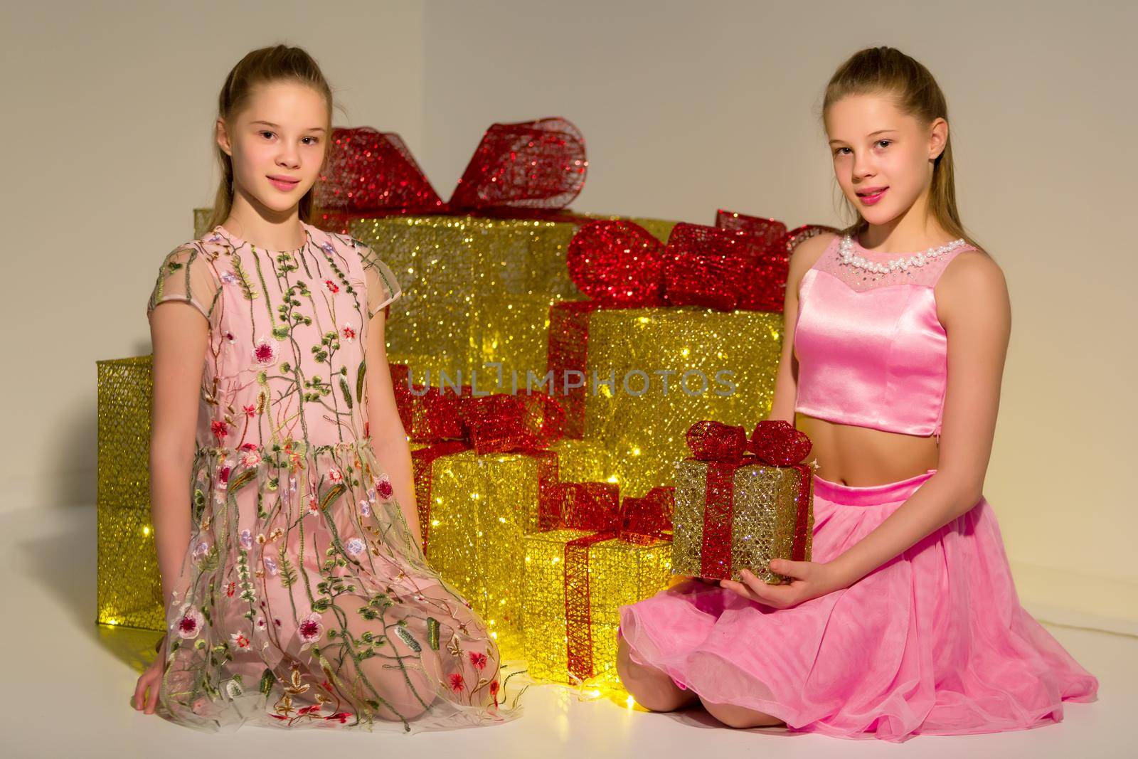 Adorable Sisters Sitting on Floor on Background Piled Gift Boxes with Red Ribbons, Portrait of Beautiful Teen Girls Celebrating Xmas and New Year Holidays, Twin Sisters Posing in Fashionable Clothes