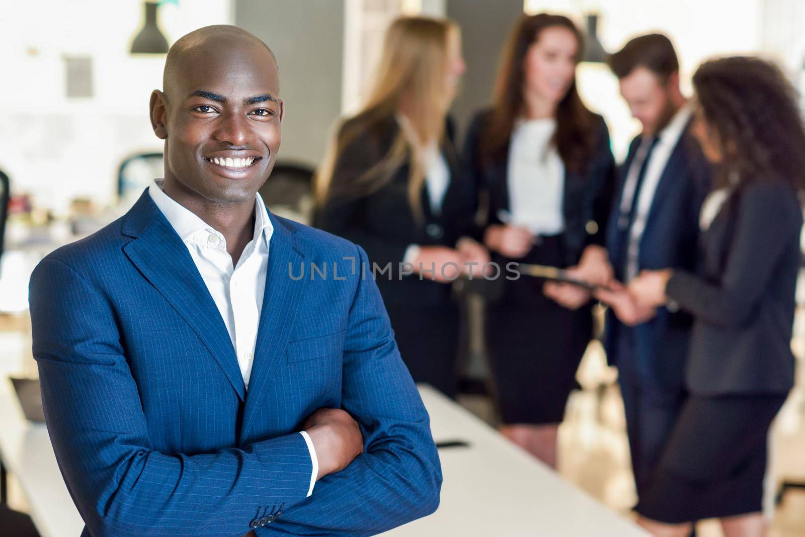 Black businessman leader looking at camera in modern office with multi-ethnic businesspeople working at the background. Teamwork concept. African man wearing blue suit.