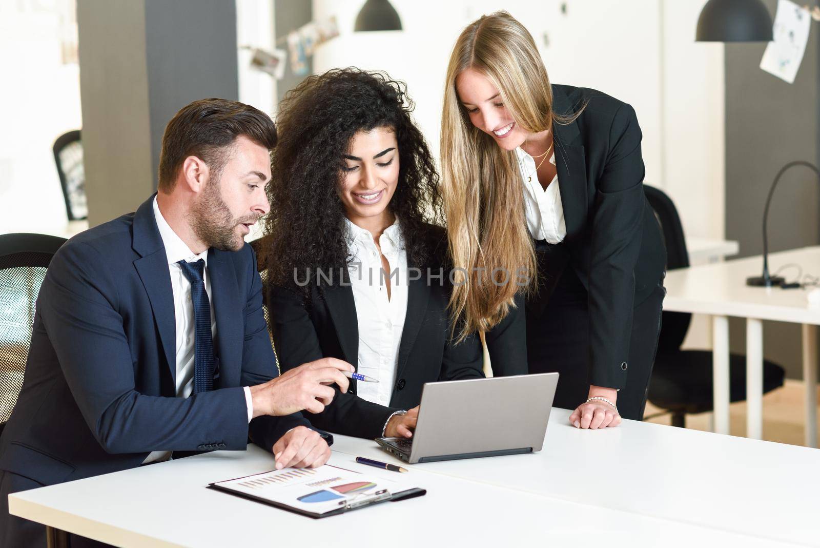 Multi-ethnic group of three businesspeople meeting in a modern office. Two women and a caucasian man wearing suit looking at a laptop computer.