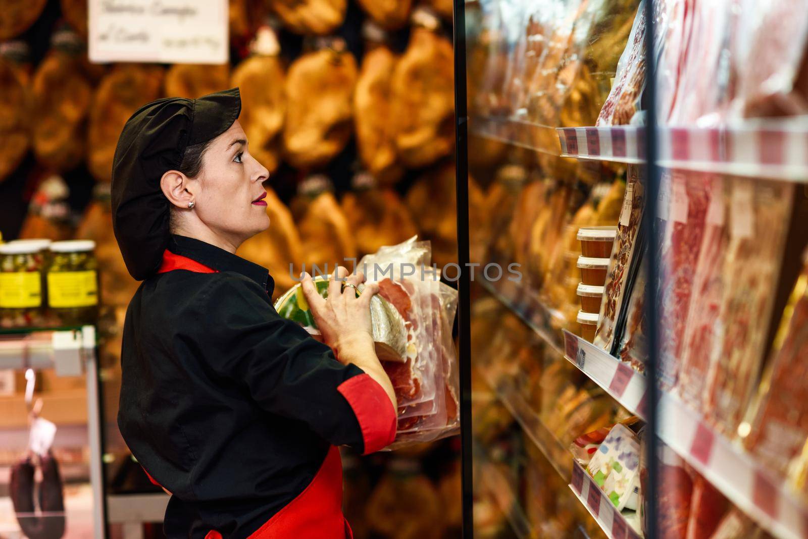 Portrait of female worker taking products in butcher shop. Refrigerated display case for sausage packages, ham and cheese