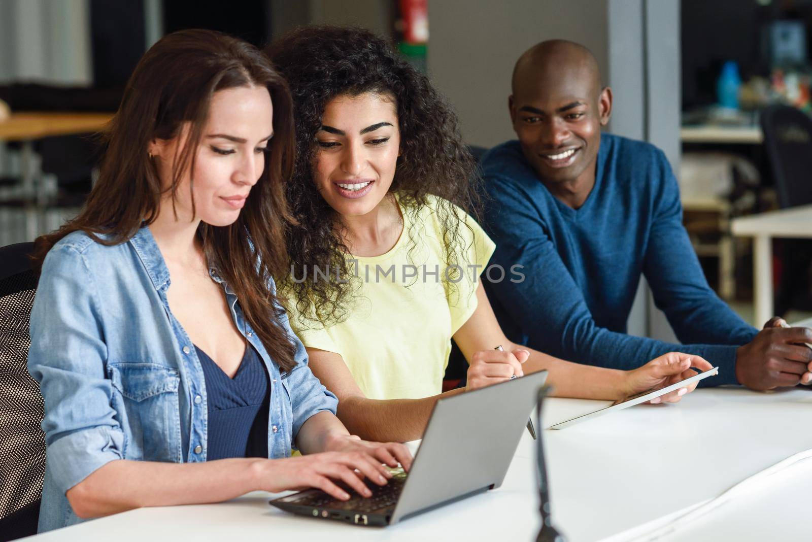 Three young people studying with laptop computer on white desk. Beautiful girls and man working toghether wearing casual clothes. Multi-ethnic group.