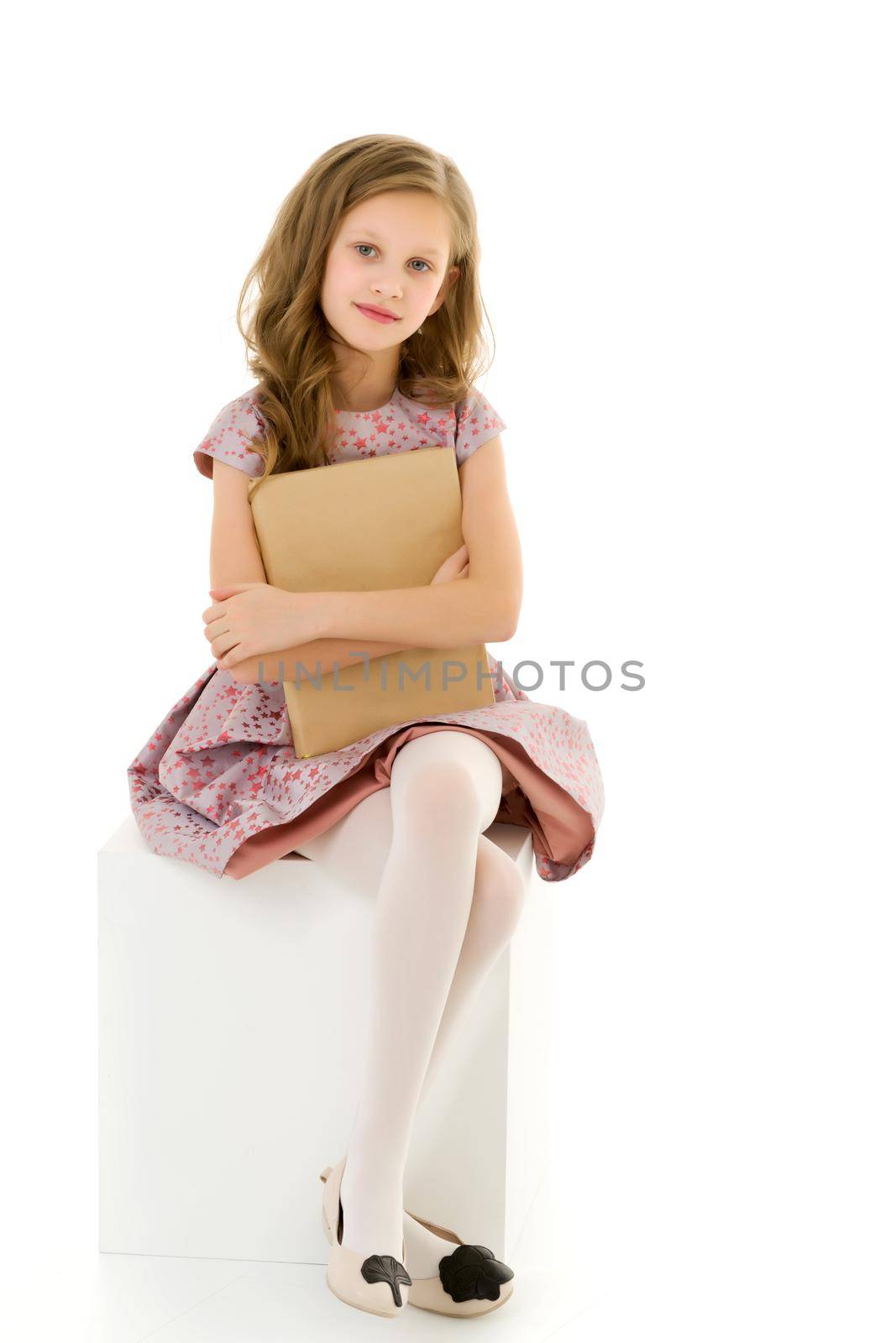 Beautiful Girl Sitting on White Cube and Hugging Book, Adorable Blonde Long Haired Girl in Stylish Clothes, Cute Elementary School Student Posing Against White Background