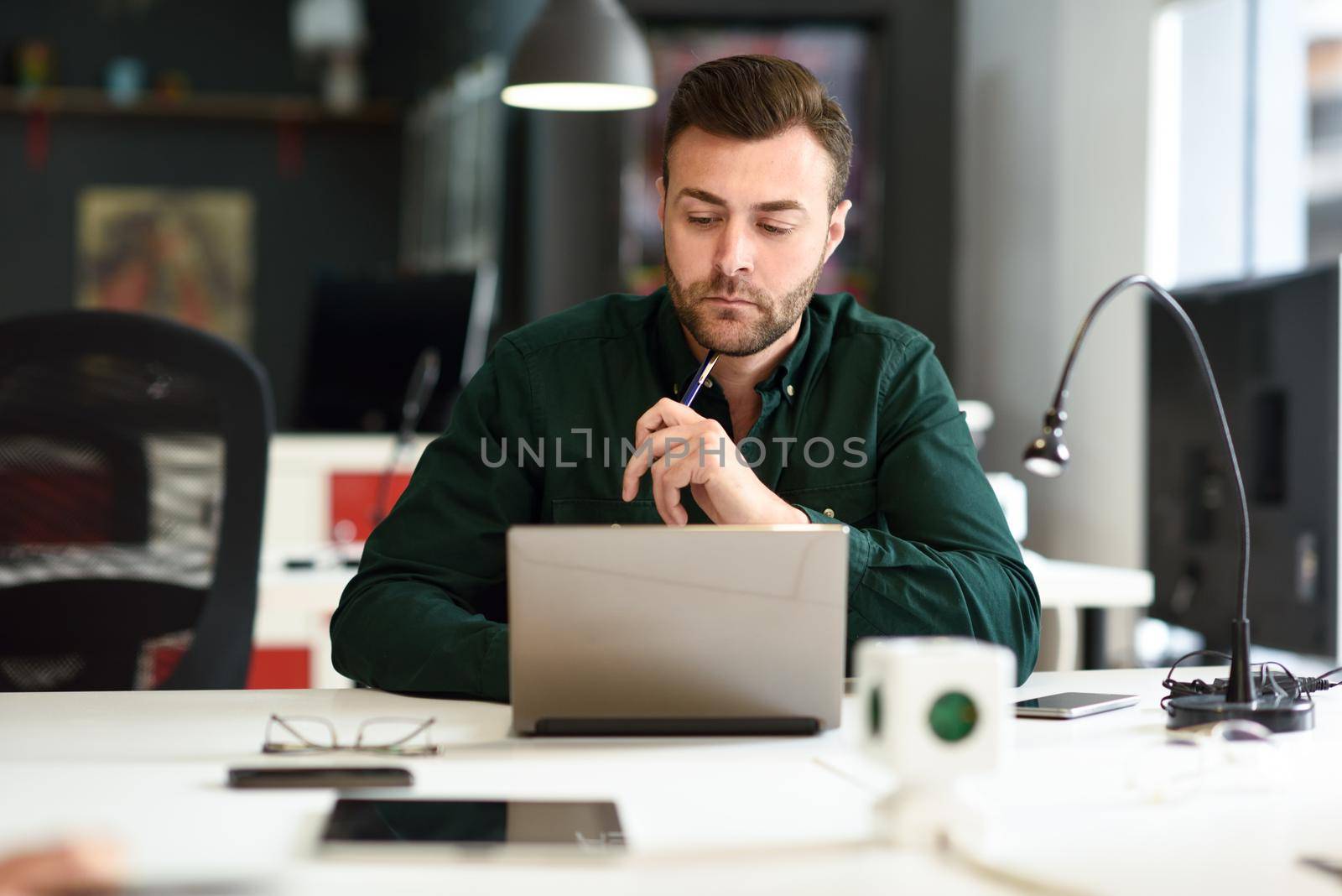 Young man studying with laptop computer on white desk. Attractive guy with beard wearing casual clothes.