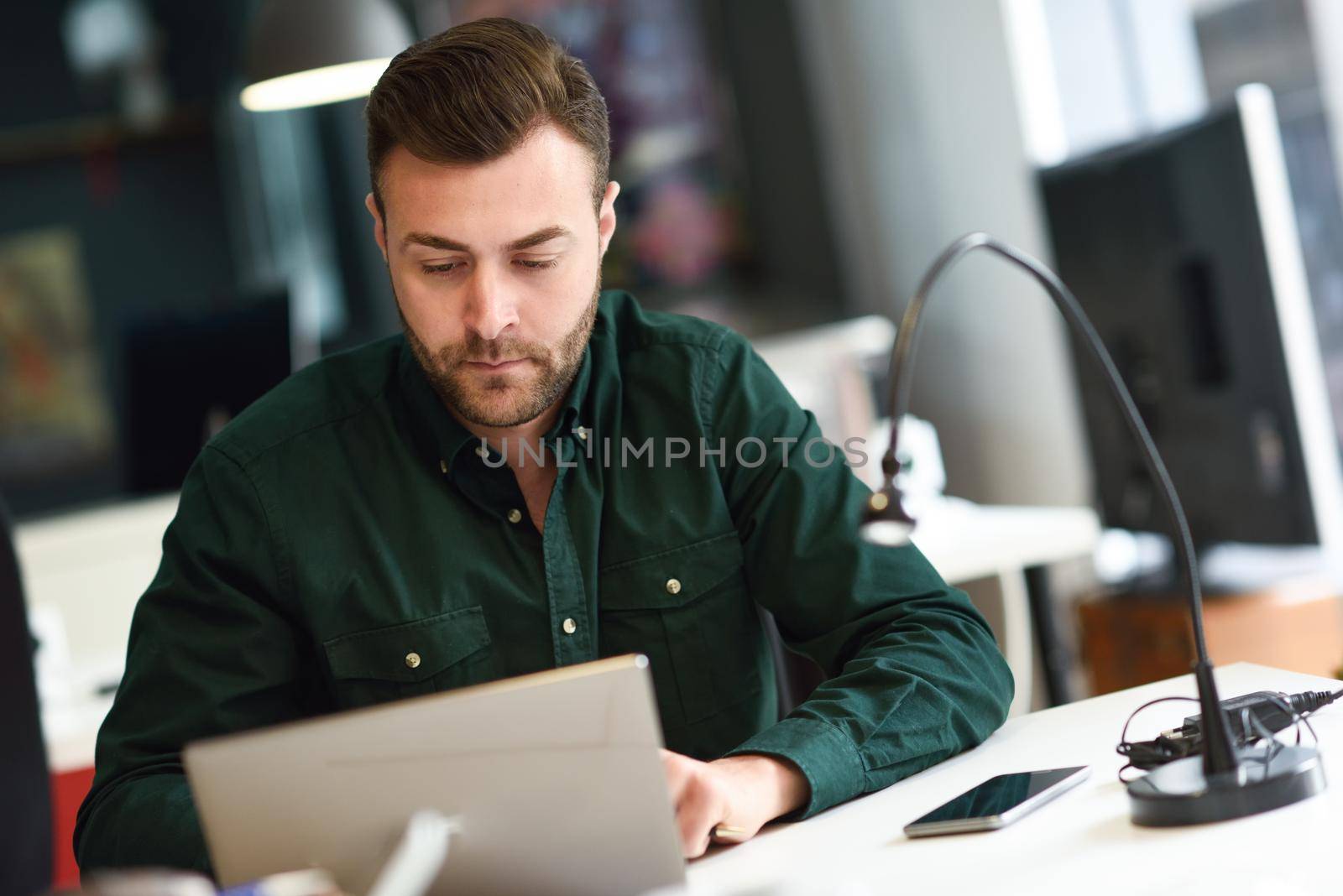 Young man studying with laptop computer on white desk. Attractive guy with beard wearing casual clothes.