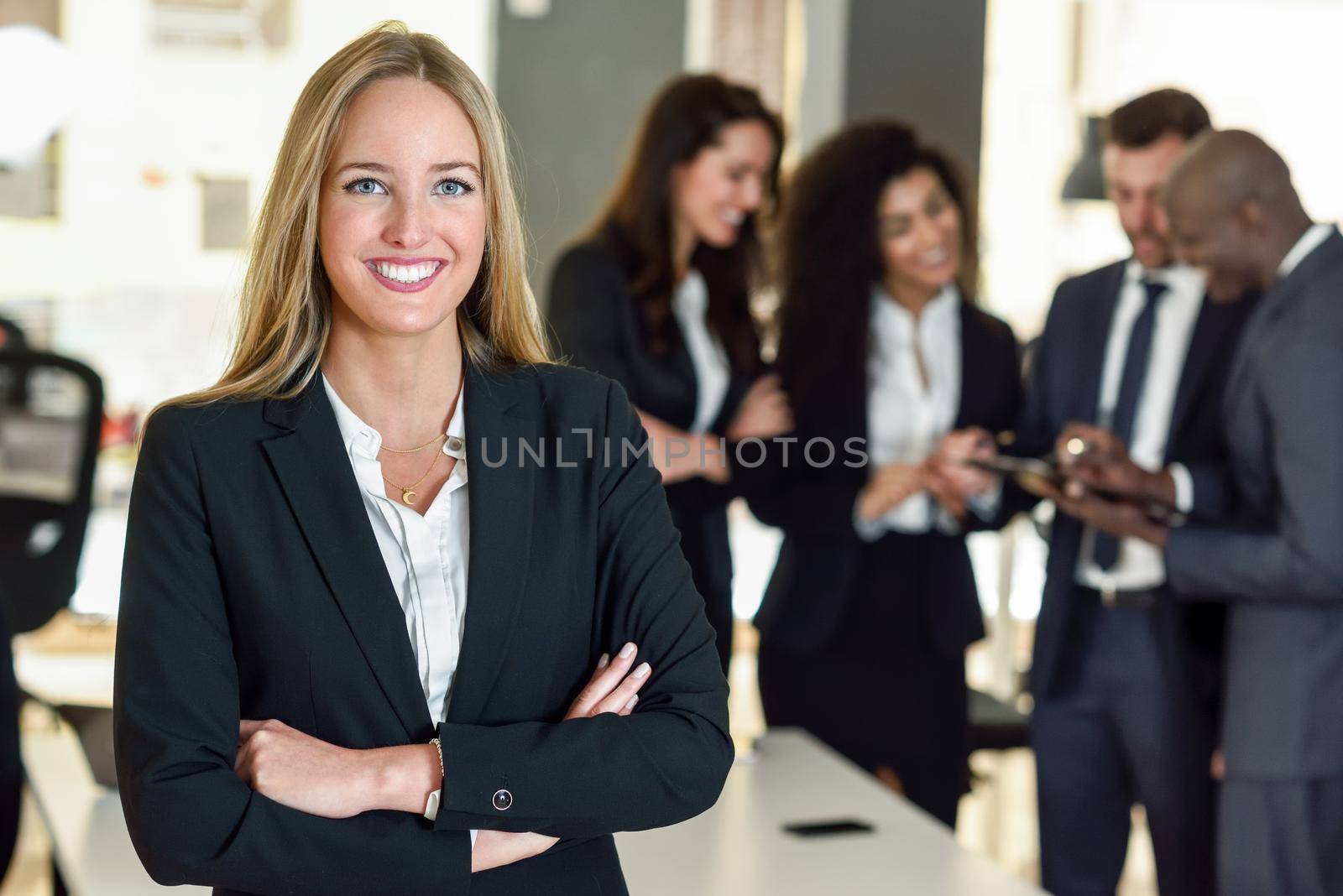 Businesswoman leader looking at camera in modern office with multi-ethnic businesspeople working at the background. Teamwork concept. Blonde caucasian woman