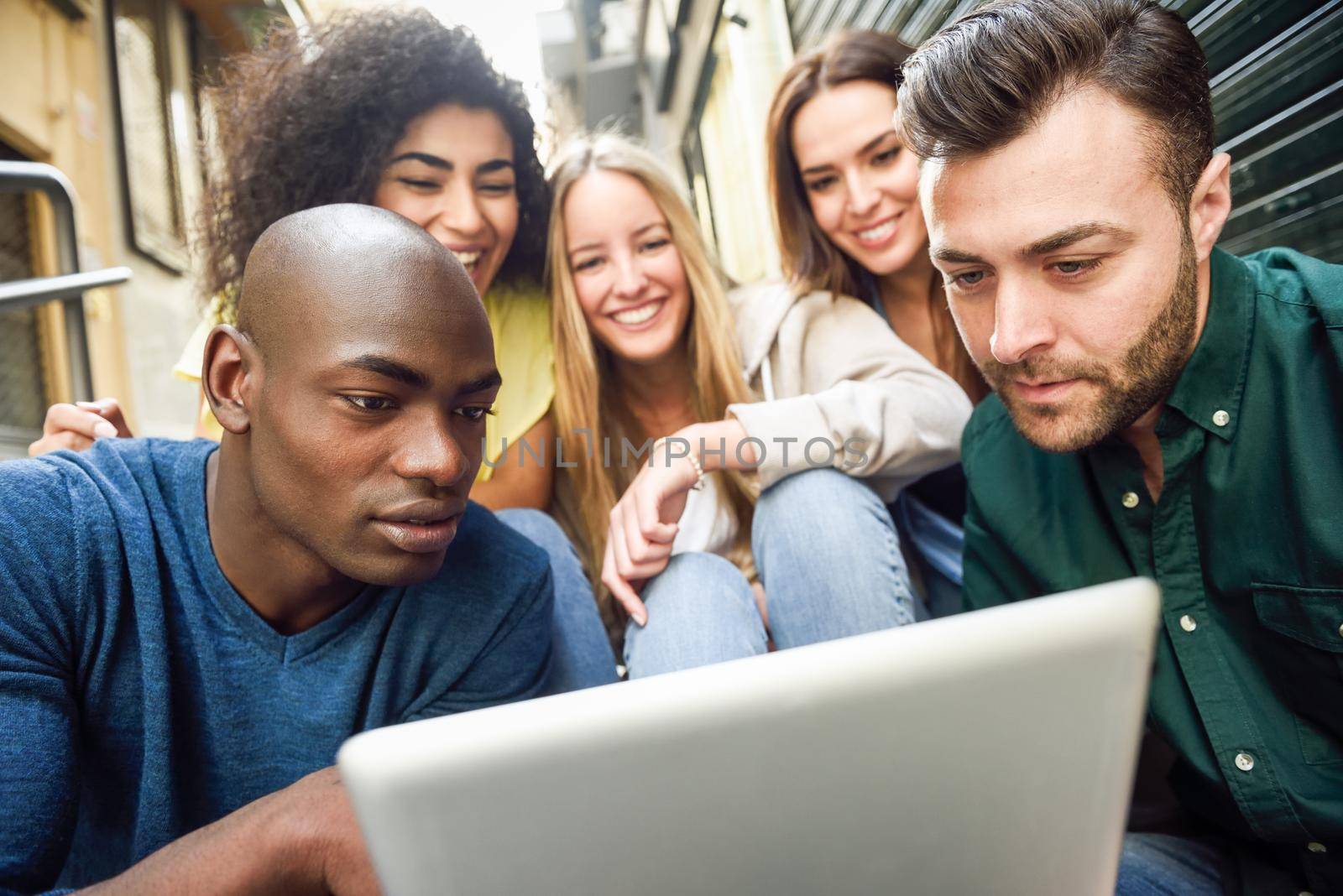 Multi-ethnic group of young people looking at a tablet computer outdoors in urban background. Group of men and woman sitting together on steps.