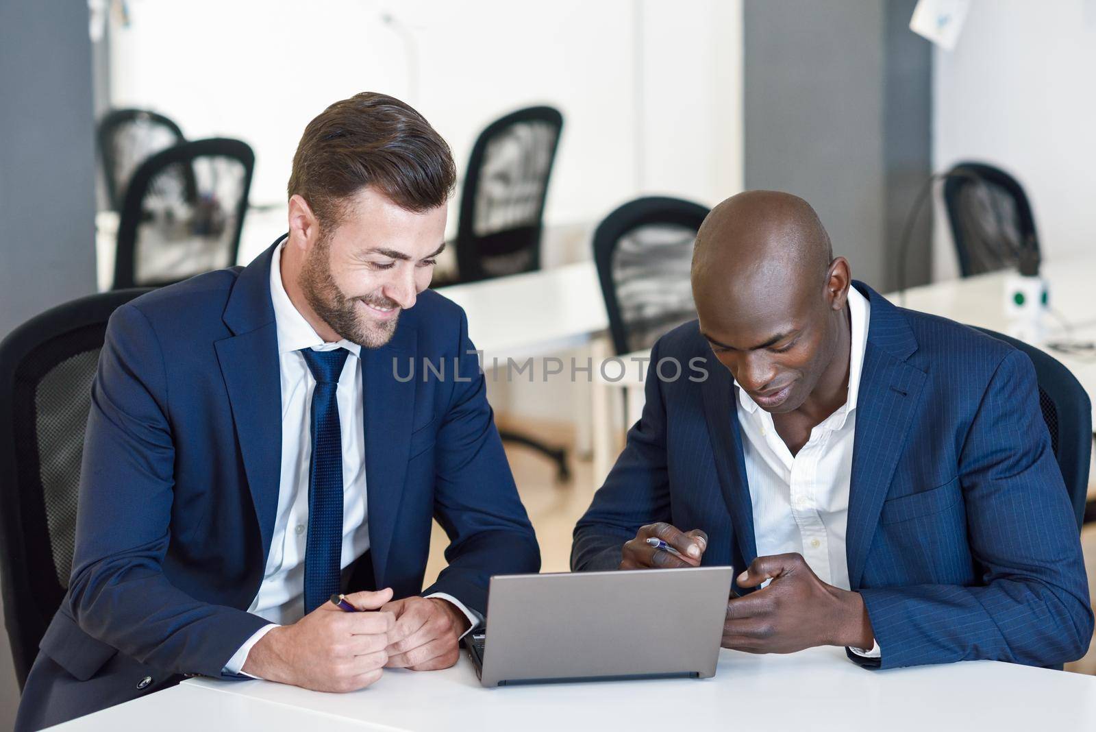 Black and caucasian smiling businessmen looking at a laptop computer. Two men wearing blue suits working in an office with white furniture