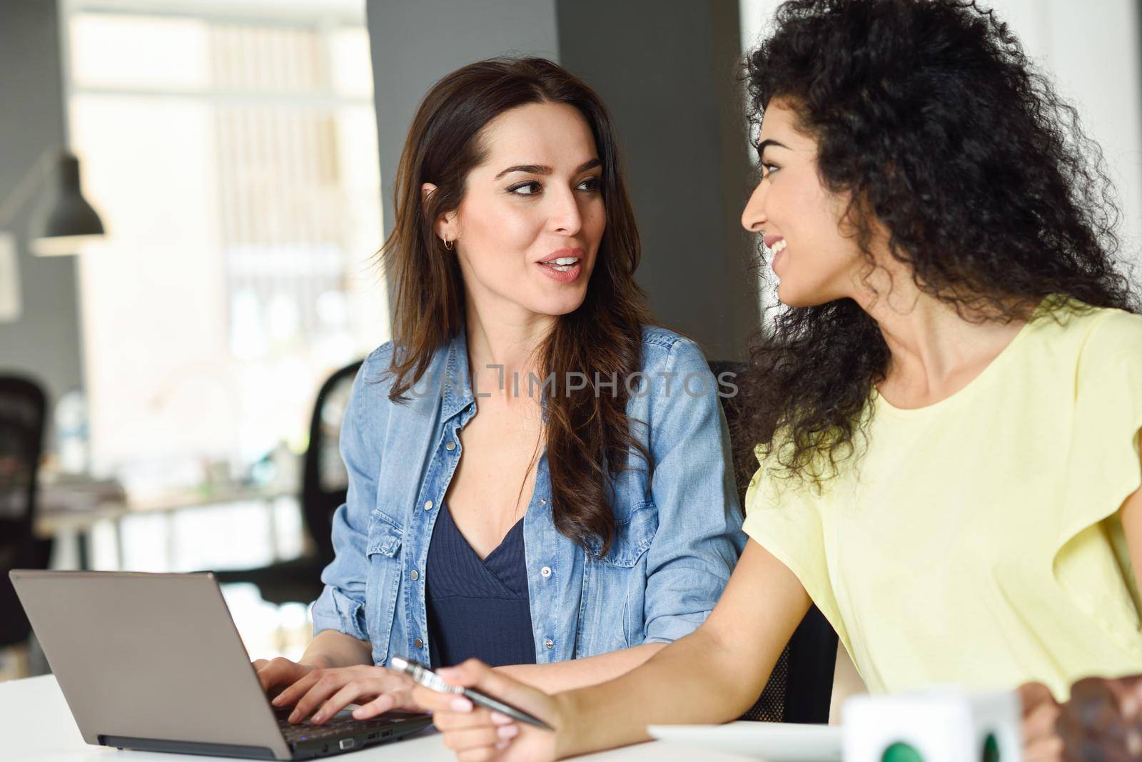 Two young women studying with a laptop computer on white desk. Beautiful girls working toghether wearing casual clothes.