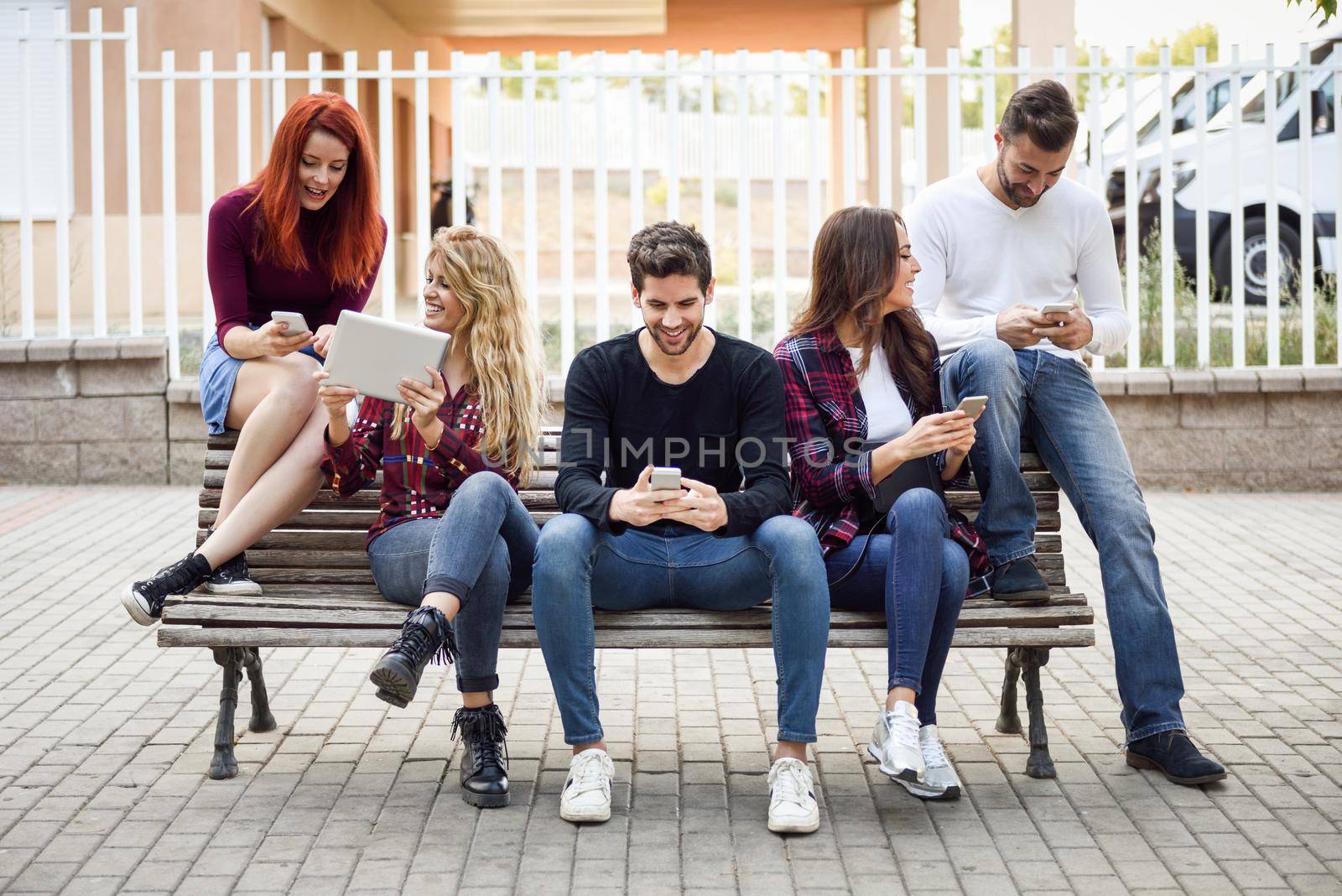 Group of young people using smartphone and tablet computers outdoors in urban background. Women and men sitting on a bench in the street wearing casual clothes.