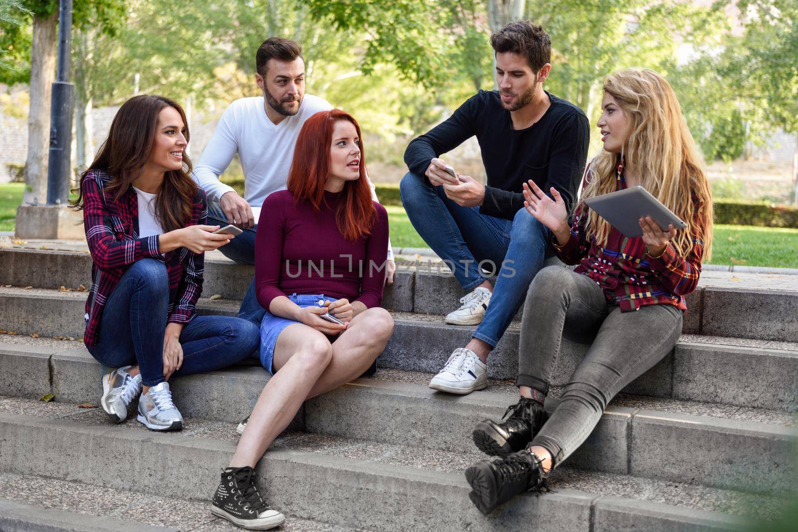 Group of young people with smartphone and tablet computers outdoors in urban background. Women and men talking on stairs in the street wearing casual clothes.