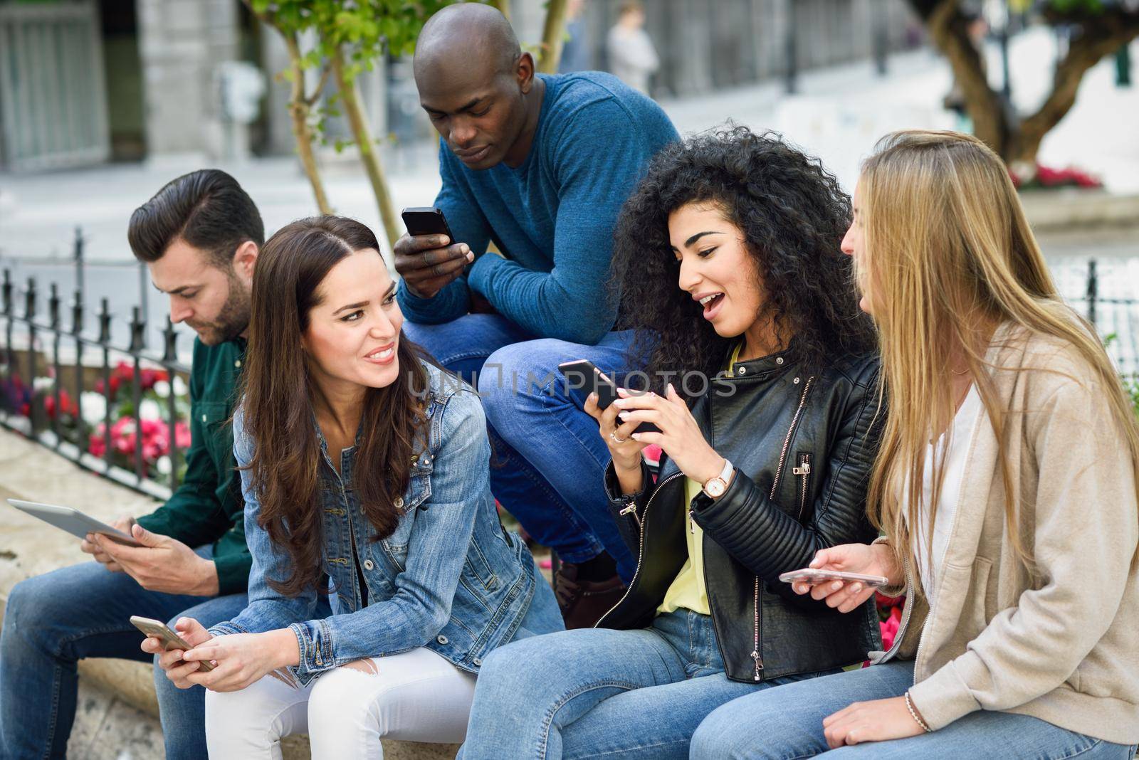 Multi-ethnic group of young people using smartphone and tablet computers outdoors in urban background. Women and men smiling and laughing in the street wearing casual clothes.