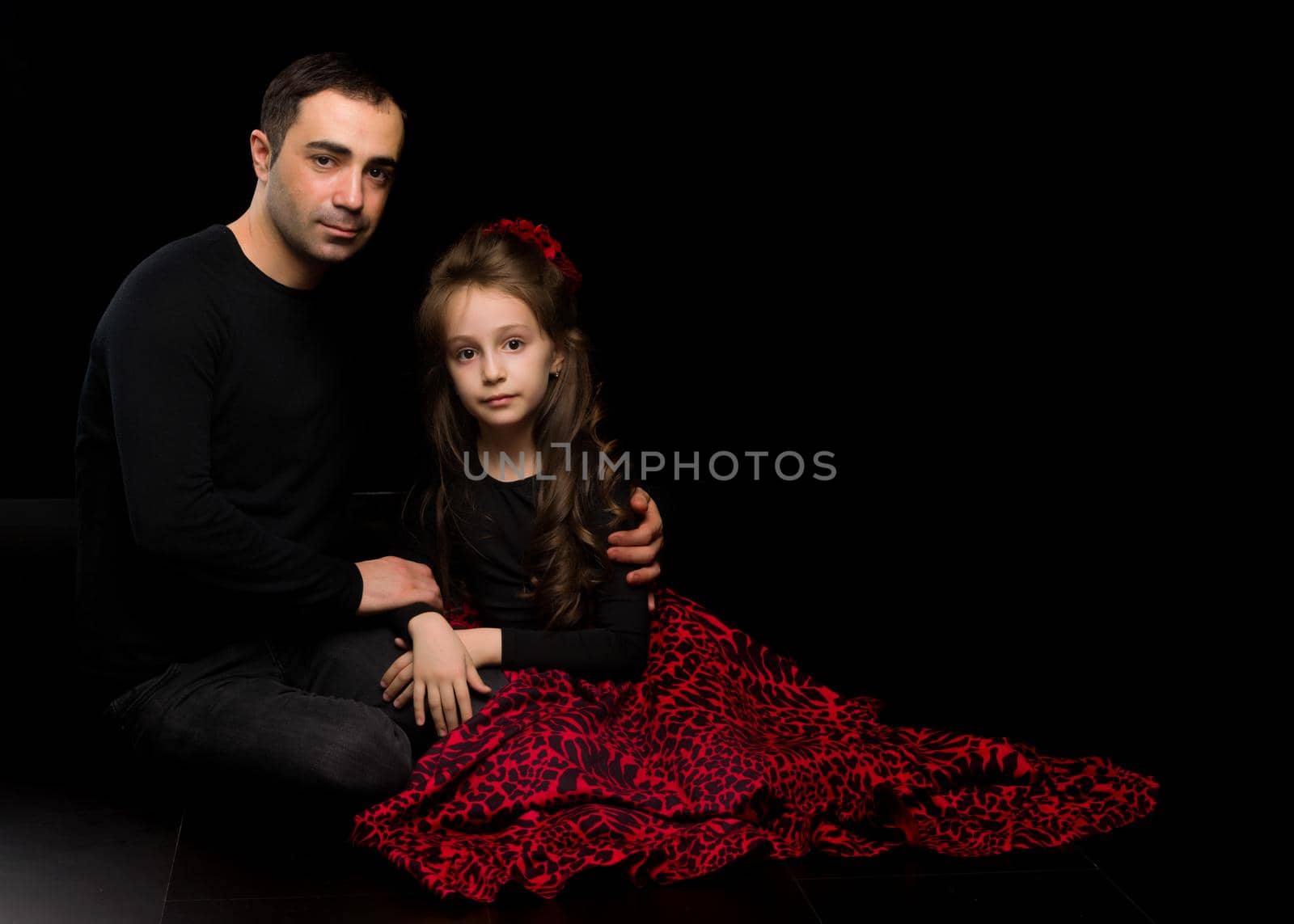 Portrait of Loving Father Hugging His Beautiful Daughter, Handsome Young Man Sitting on the Floor with his Adorable Girl, Happy Family Sitting Against Black Studio Background