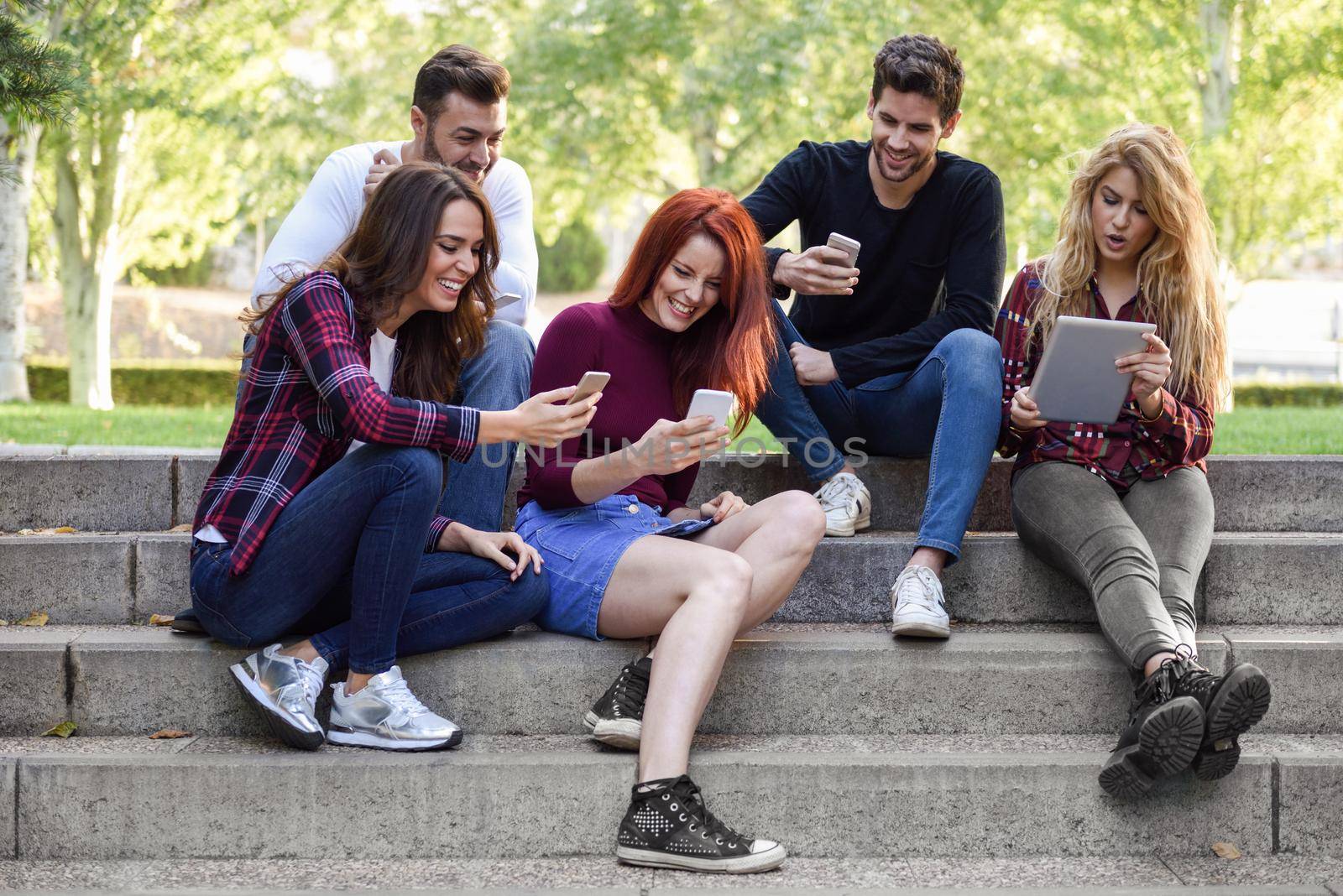 Group of young people using smartphone and tablet computers outdoors in urban background. Women and men sitting on stairs in the street wearing casual clothes.