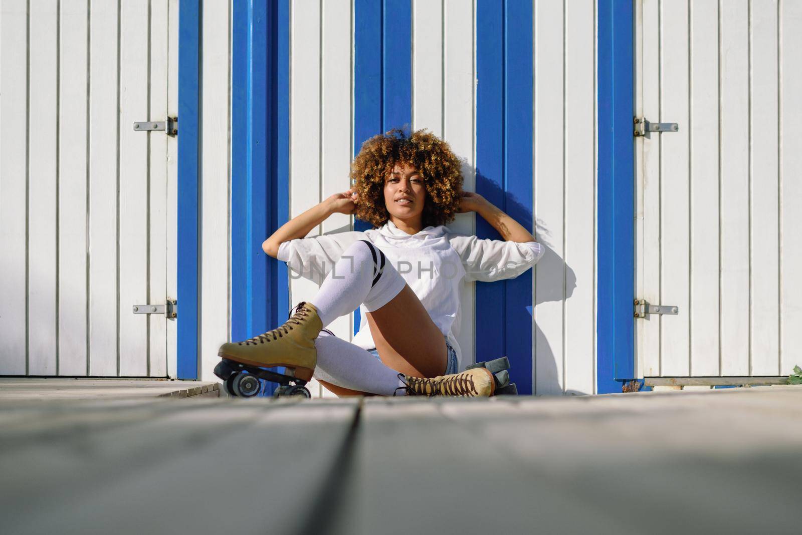 Young black woman on roller skates sitting near a beach hut. Girl with afro hairstyle rollerblading on sunny promenade.