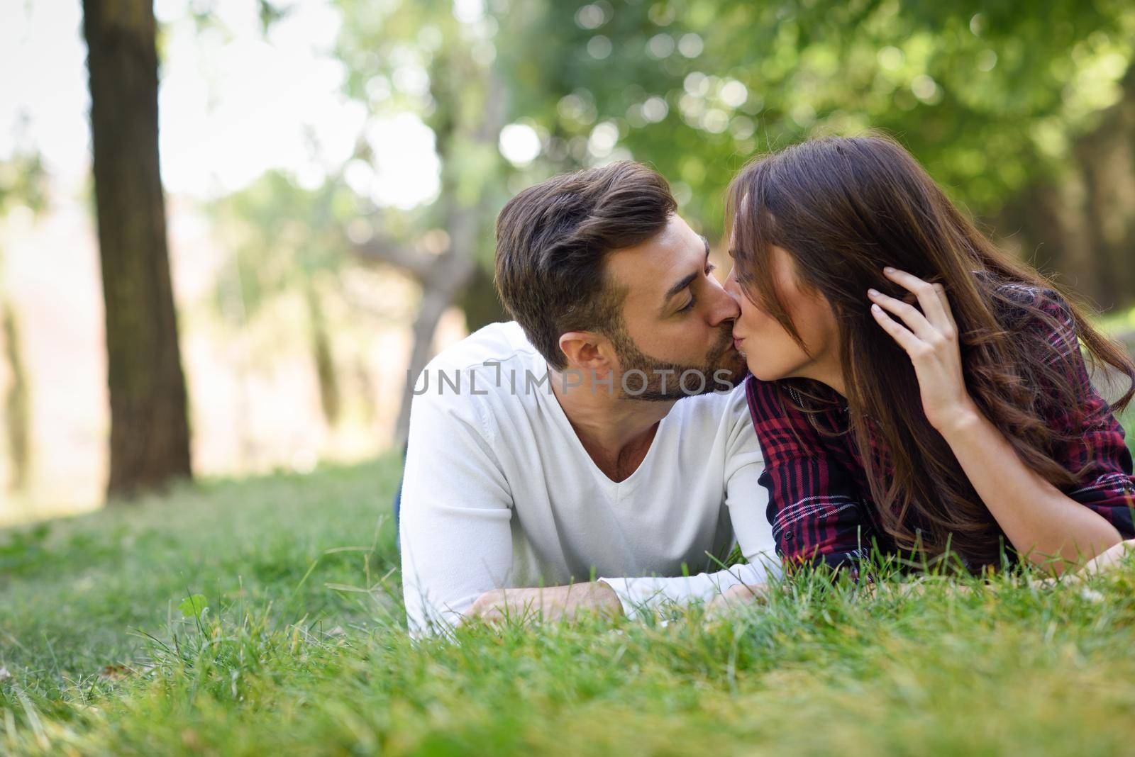 Beautiful young couple kissing on grass in an urban park. Caucasian man and woman wearing casual clothes.