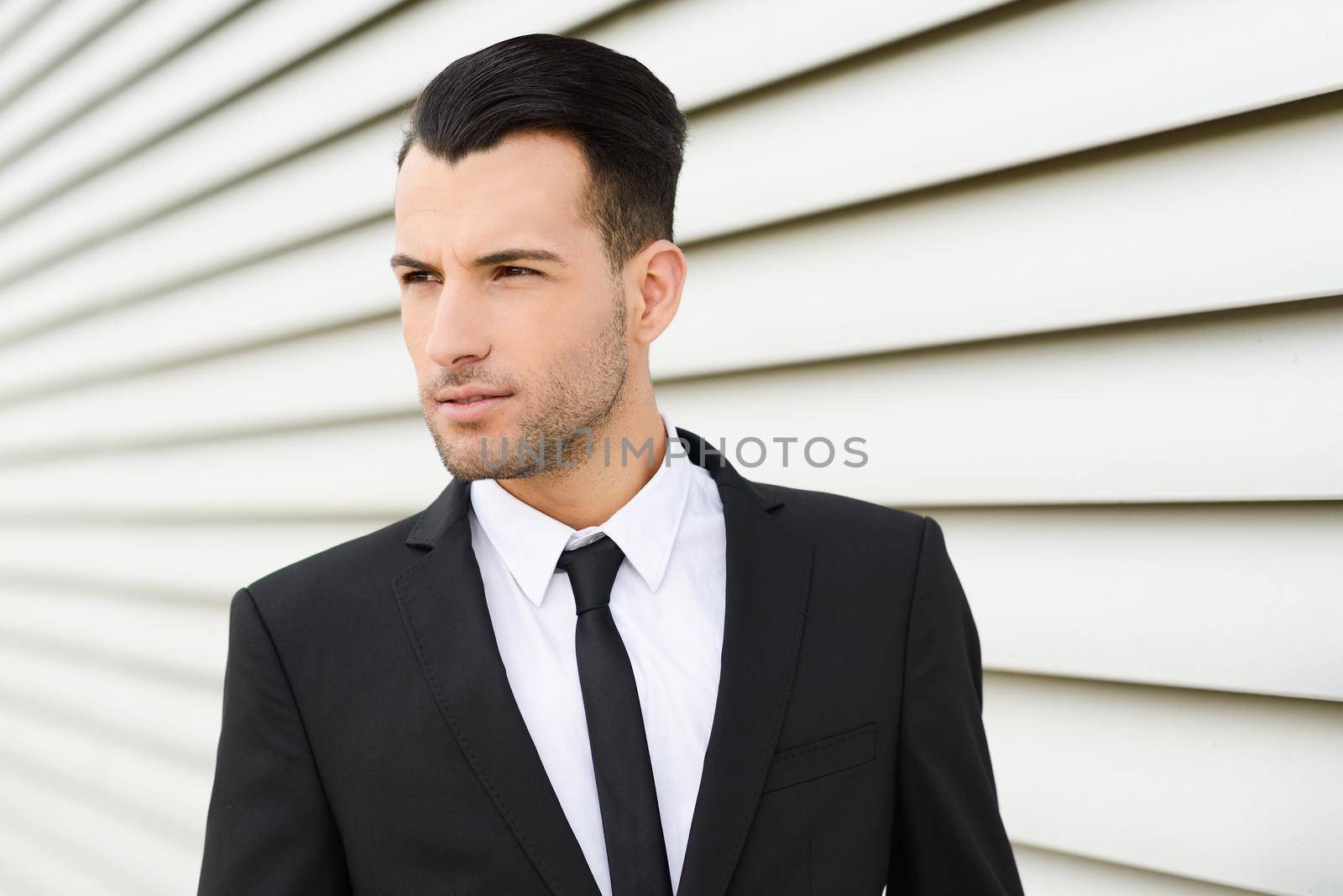 Portrait of a young businessman near a office building wearing black suit and tie with modern haircut