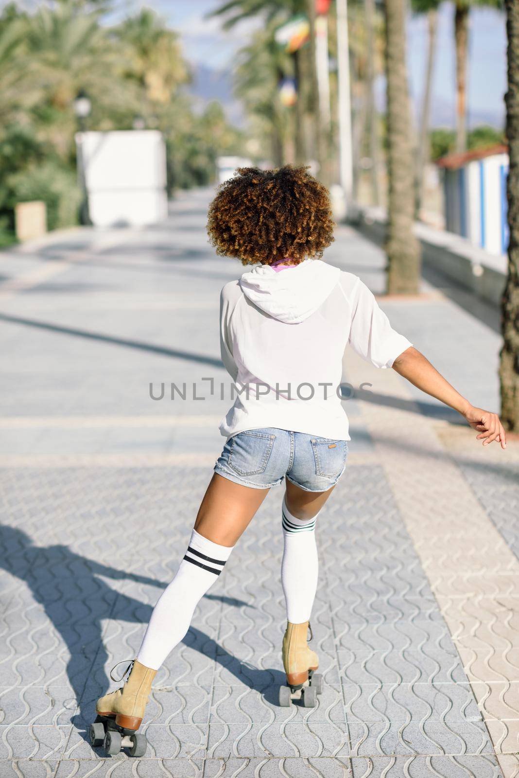 Rear view of black woman on roller skates riding outdoors on beach promenade with palm trees. Girl afro hairstyle rollerblading on sunny day.
