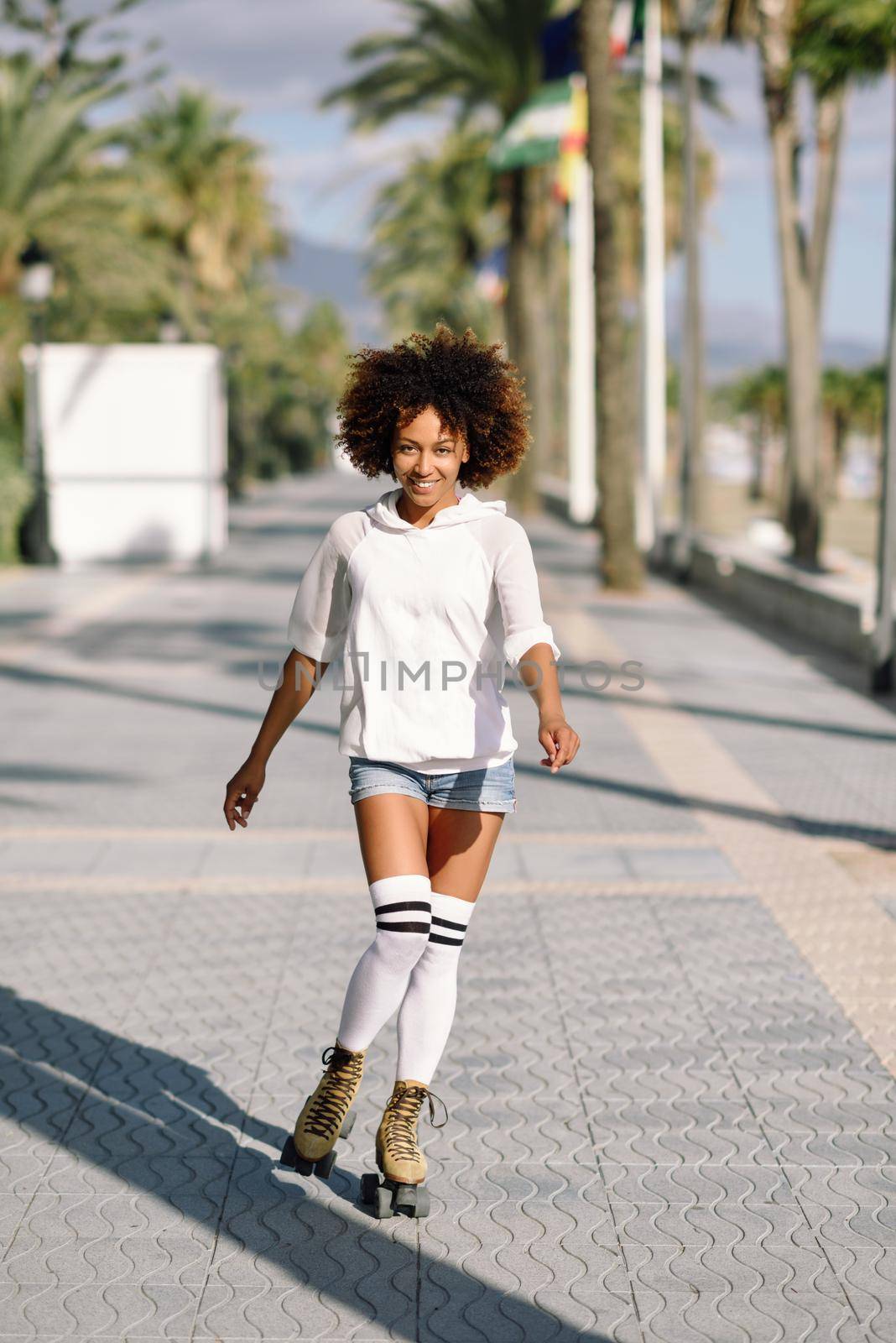 Smiling black woman on roller skates riding outdoors on beach promenade with palm trees. Smiling girl with afro hairstyle rollerblading on sunny day.