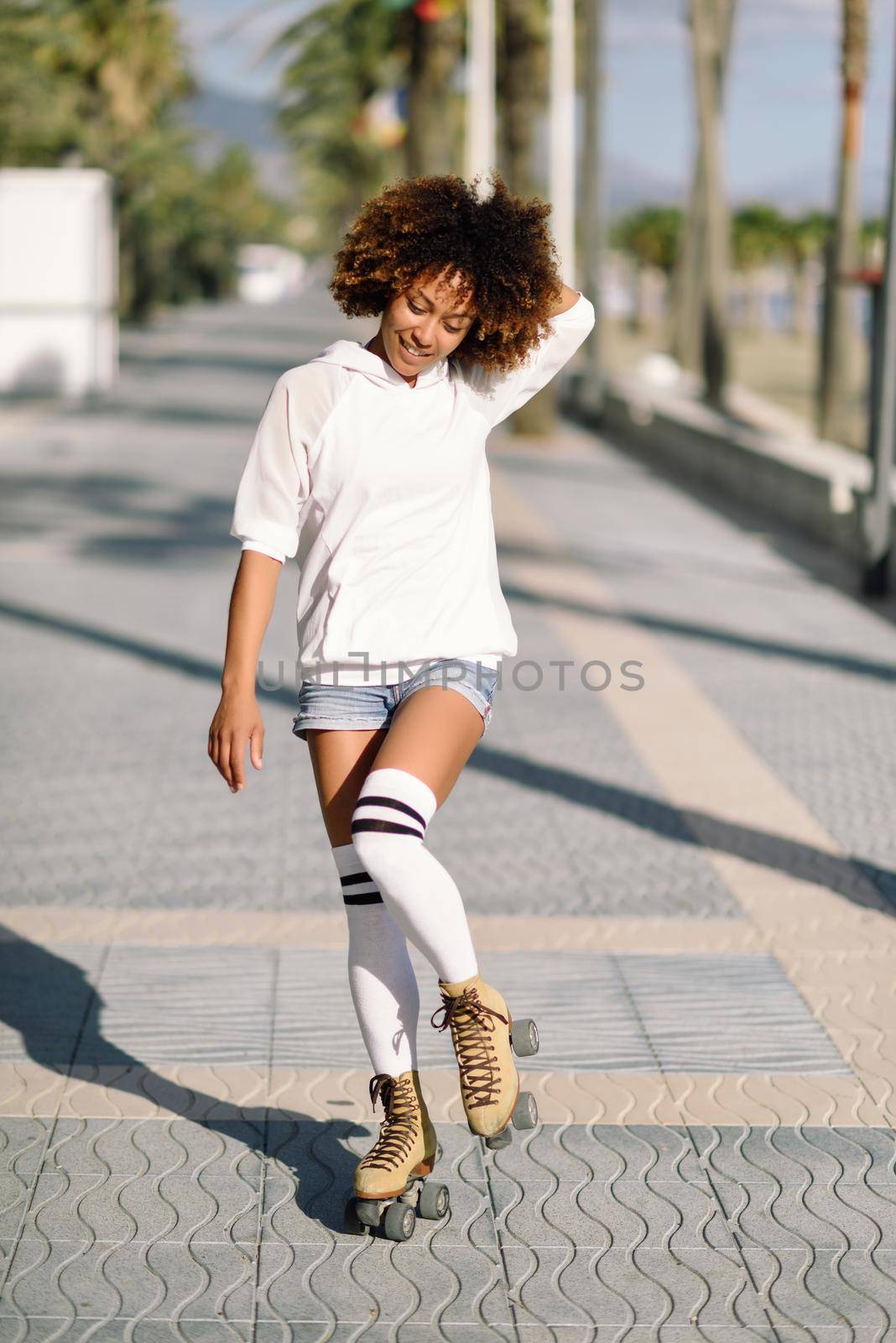 Smiling black woman on roller skates riding outdoors on beach promenade with palm trees. Smiling girl with afro hairstyle rollerblading on sunny day.