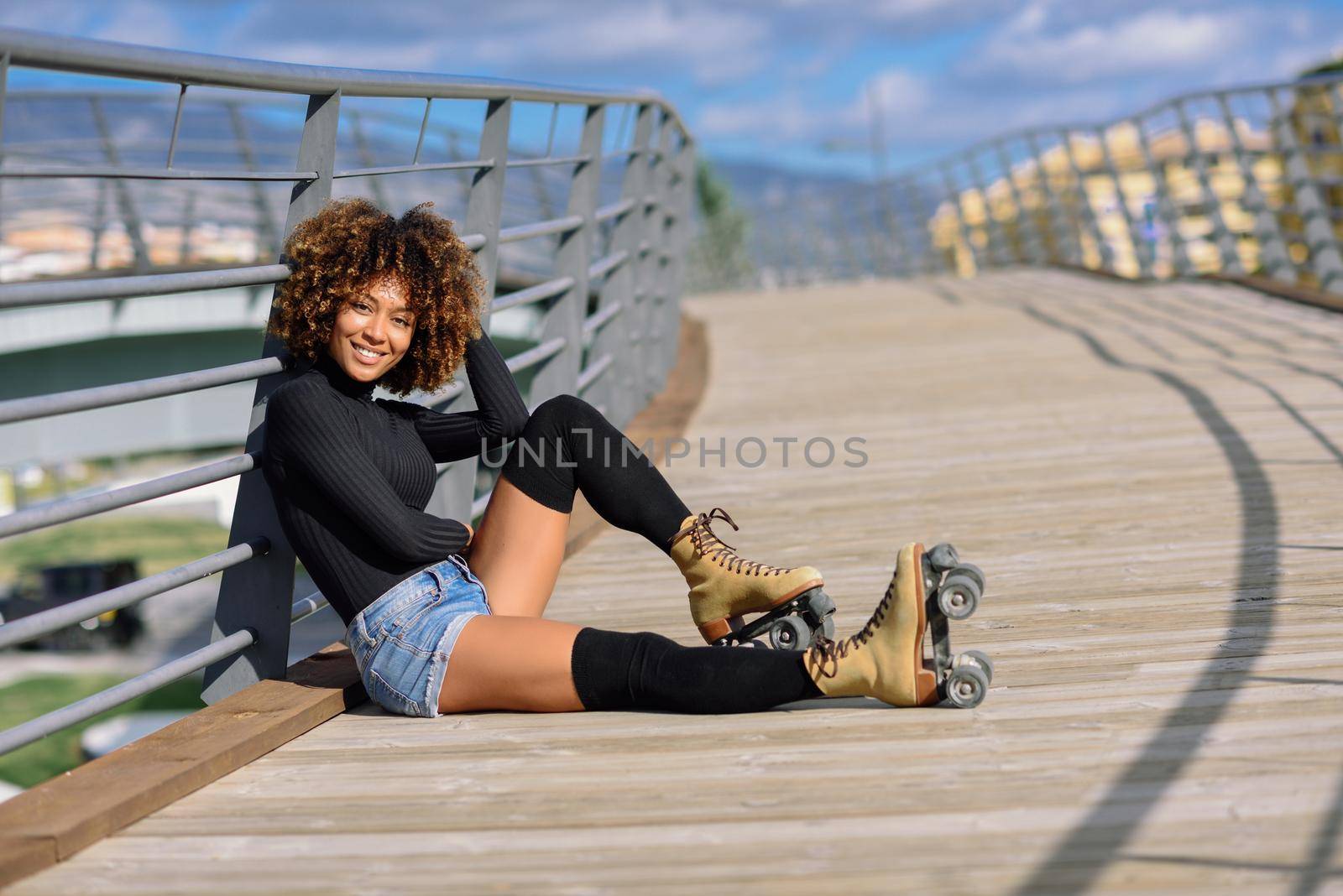 Black woman, afro hairstyle, on roller skates sitting outdoors on urban bridge. Smiling young female with beautiful clouds at the background.