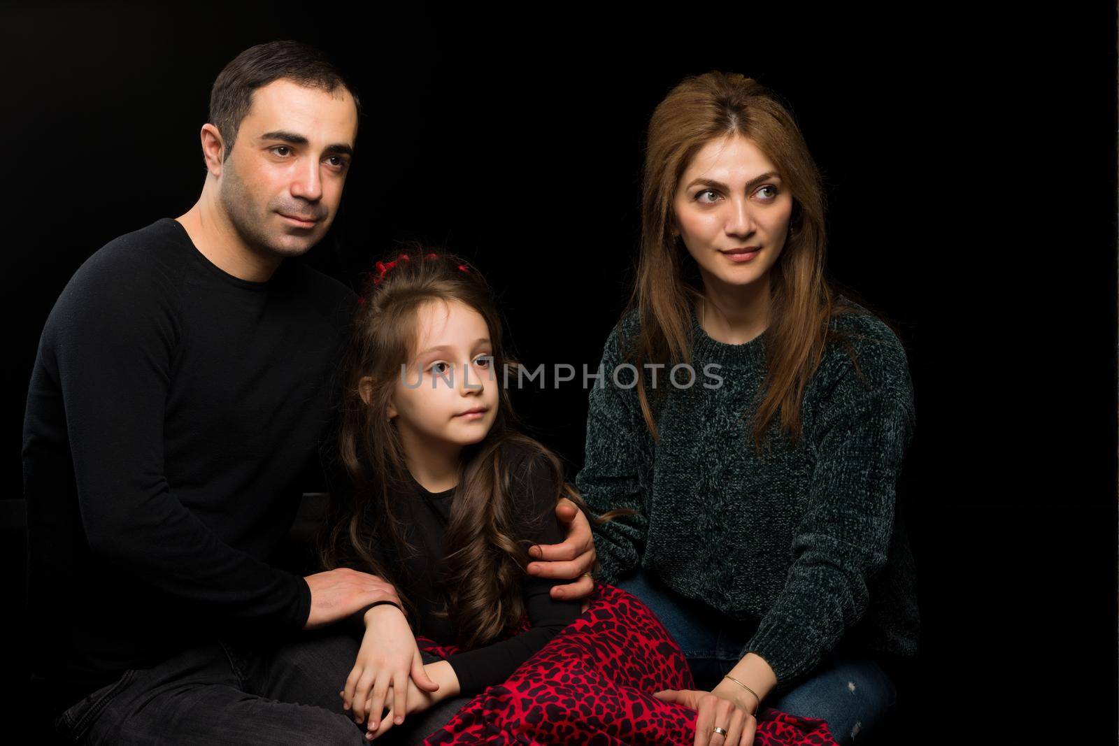 Loving Parents Hugging their Beautiful Daughter, Mother, Father and Adorable Girl Sitting on the Floor and Looking to the Side, Happy Family Posing Against Black Studio Background