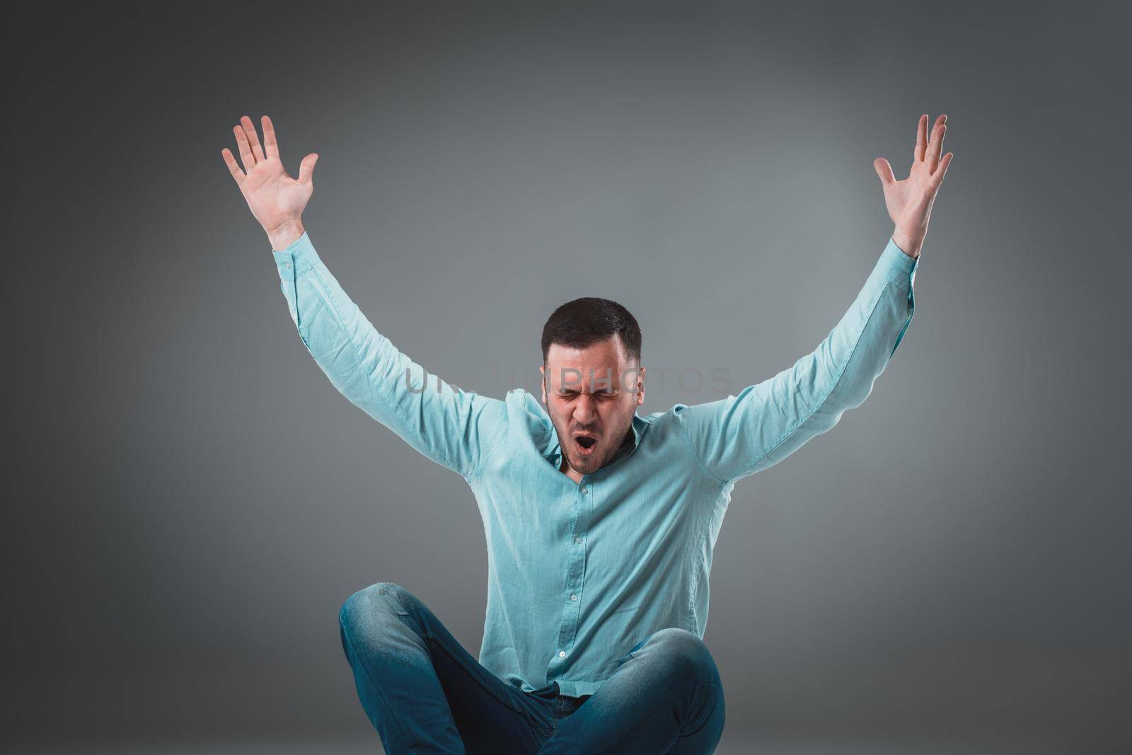 Casual young man sitting on the floor with his legs crossed and cheering with his hands in the air while screaming. Isolated on gray background
