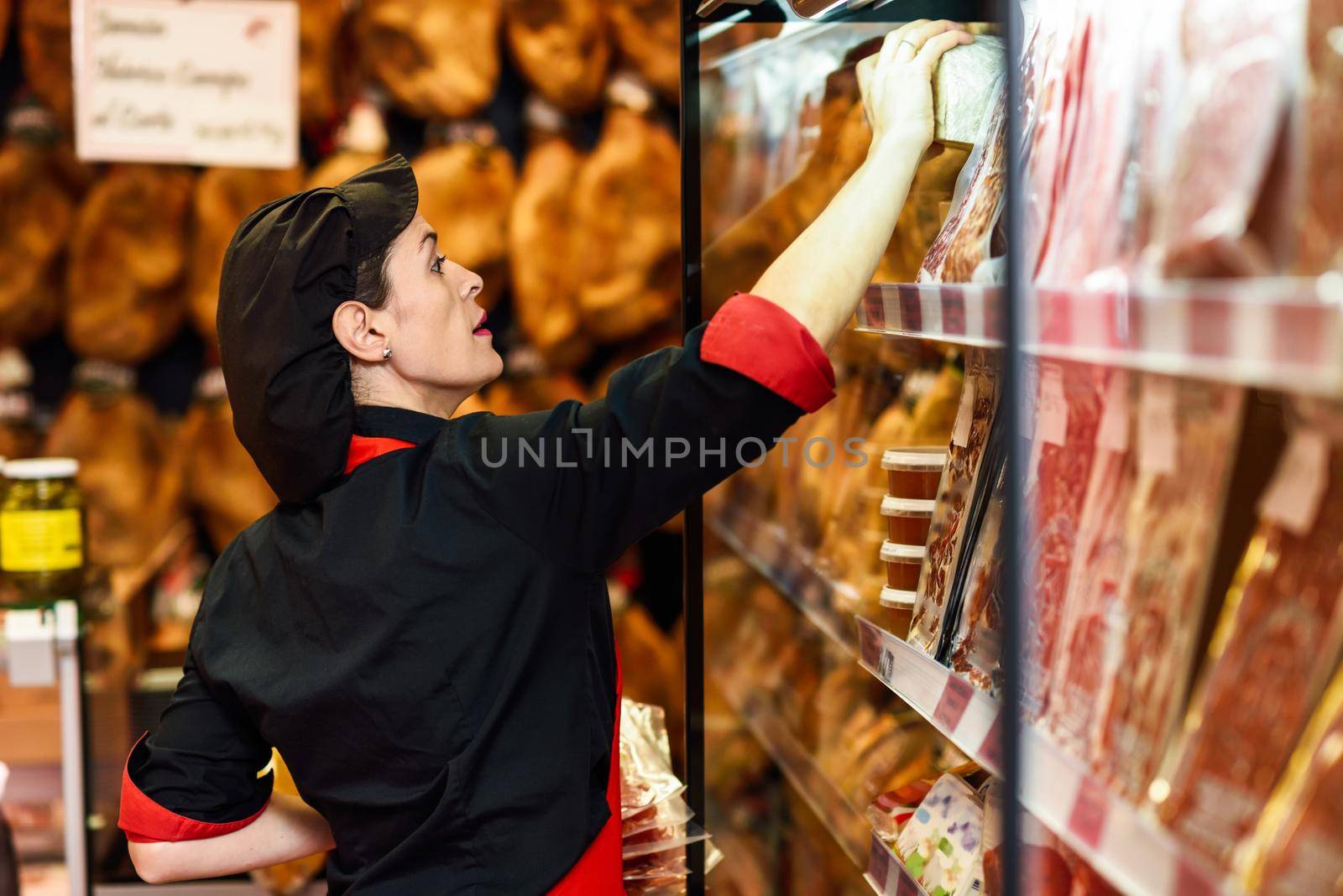 Portrait of female worker taking products in butcher shop. Refrigerated display case for sausage packages, ham and cheese