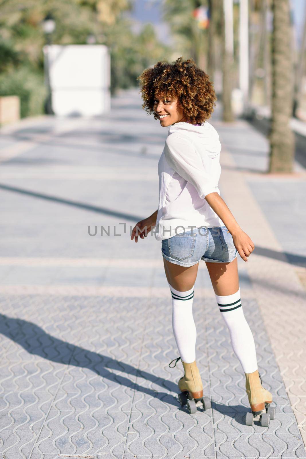 Smiling black woman on roller skates riding outdoors on beach promenade with palm trees. Smiling girl with afro hairstyle rollerblading on sunny day.